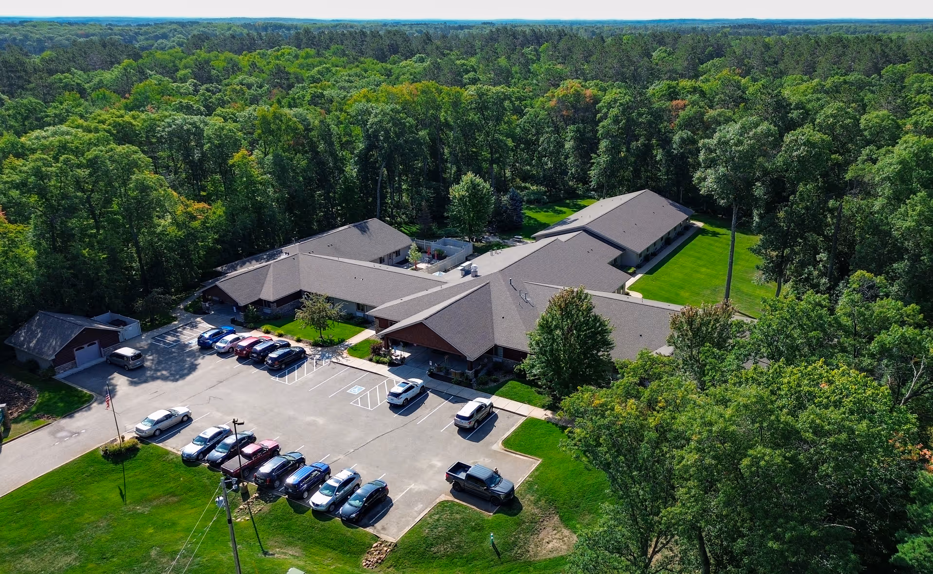 Aerial view of Golden Horizons of Crosslake facility surrounded by dense green trees. The building has multiple connected sections with gray roofs and a parking lot in front with several parked cars. The area is lush with green grass and trees extending into the horizon.