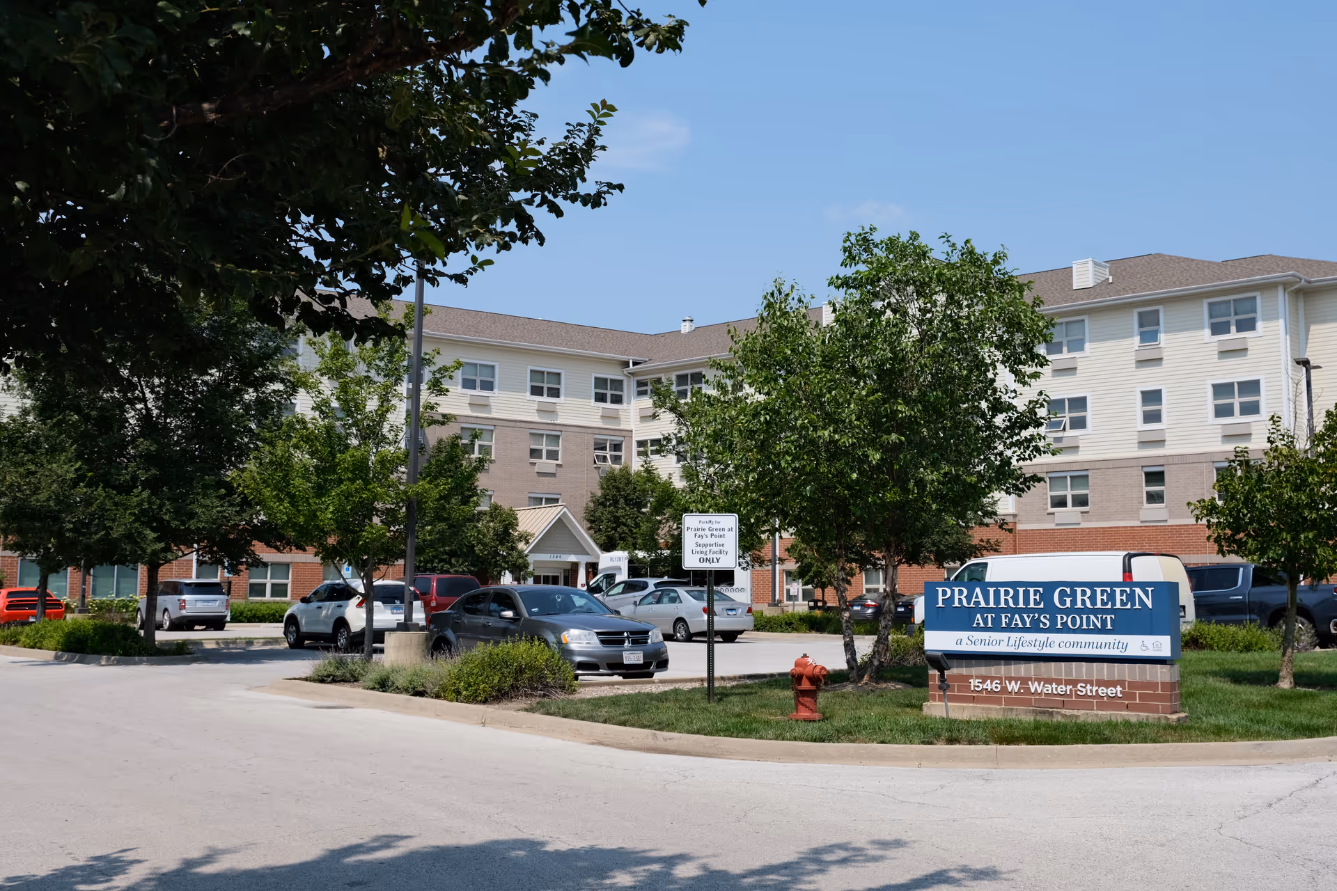 Exterior view of Prairie Green at Fay's Point senior living community building with a parking lot in front, several cars parked, trees, and a blue sign displaying the facility name and address.