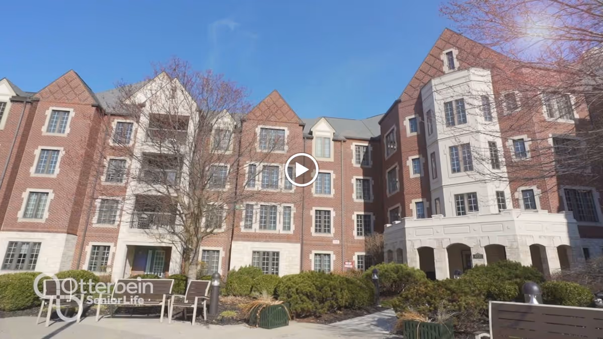 Exterior view of a multi-story senior living community building with red brick and white stone accents, surrounded by shrubs and outdoor seating under a clear blue sky.