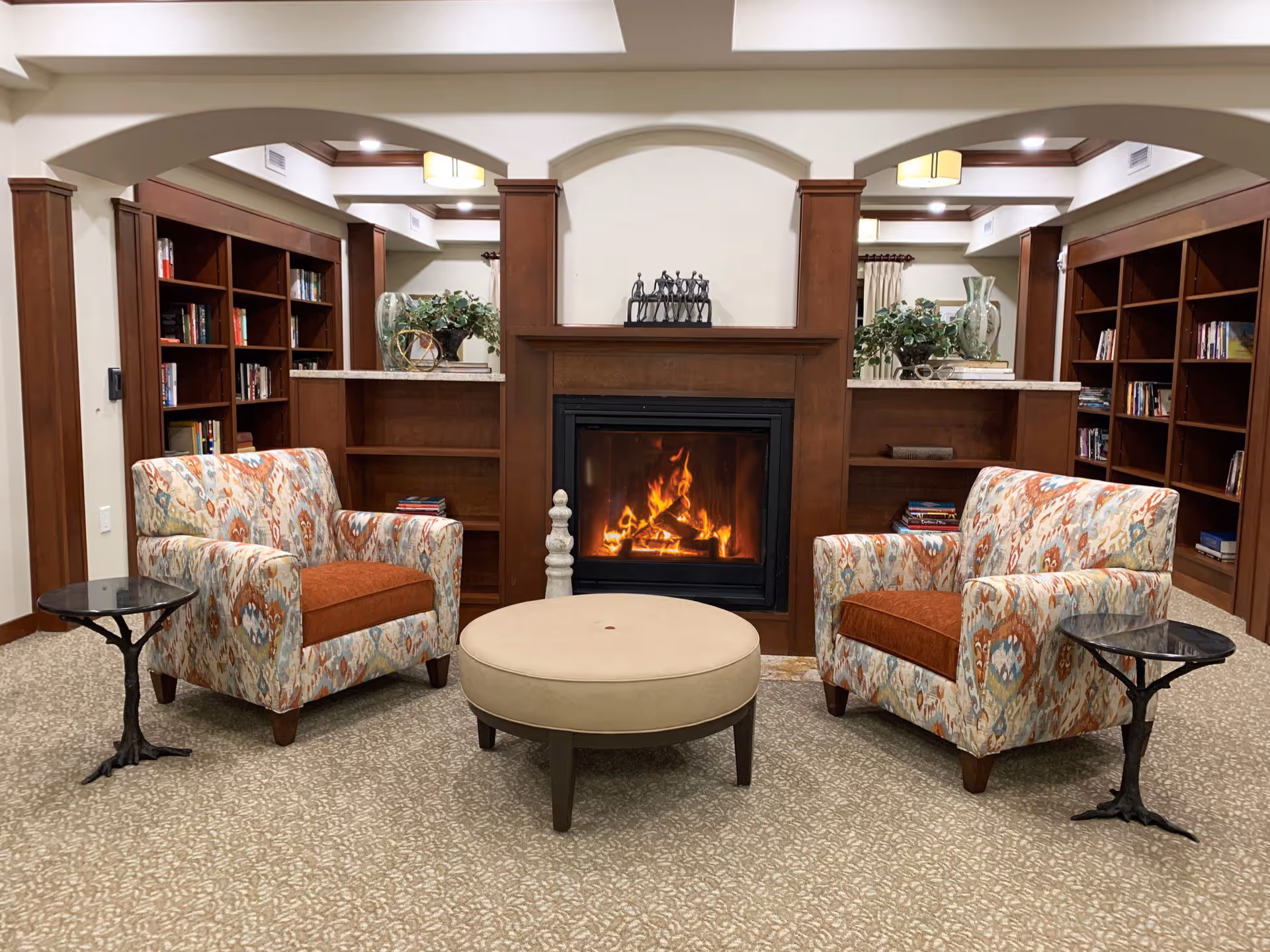 A cozy living room area with two patterned armchairs featuring orange cushions, positioned on either side of a round beige ottoman. Between the chairs are two small black side tables with decorative bases. In the background, there is a wooden fireplace with a fire burning, flanked by built-in wooden bookshelves filled with books and decorative items. The room has a beige carpet and cream-colored walls with wooden trim and arches.