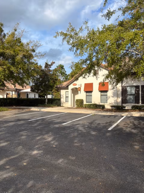 Exterior view of a single-story facility with a parking lot and trees, orange window awnings.