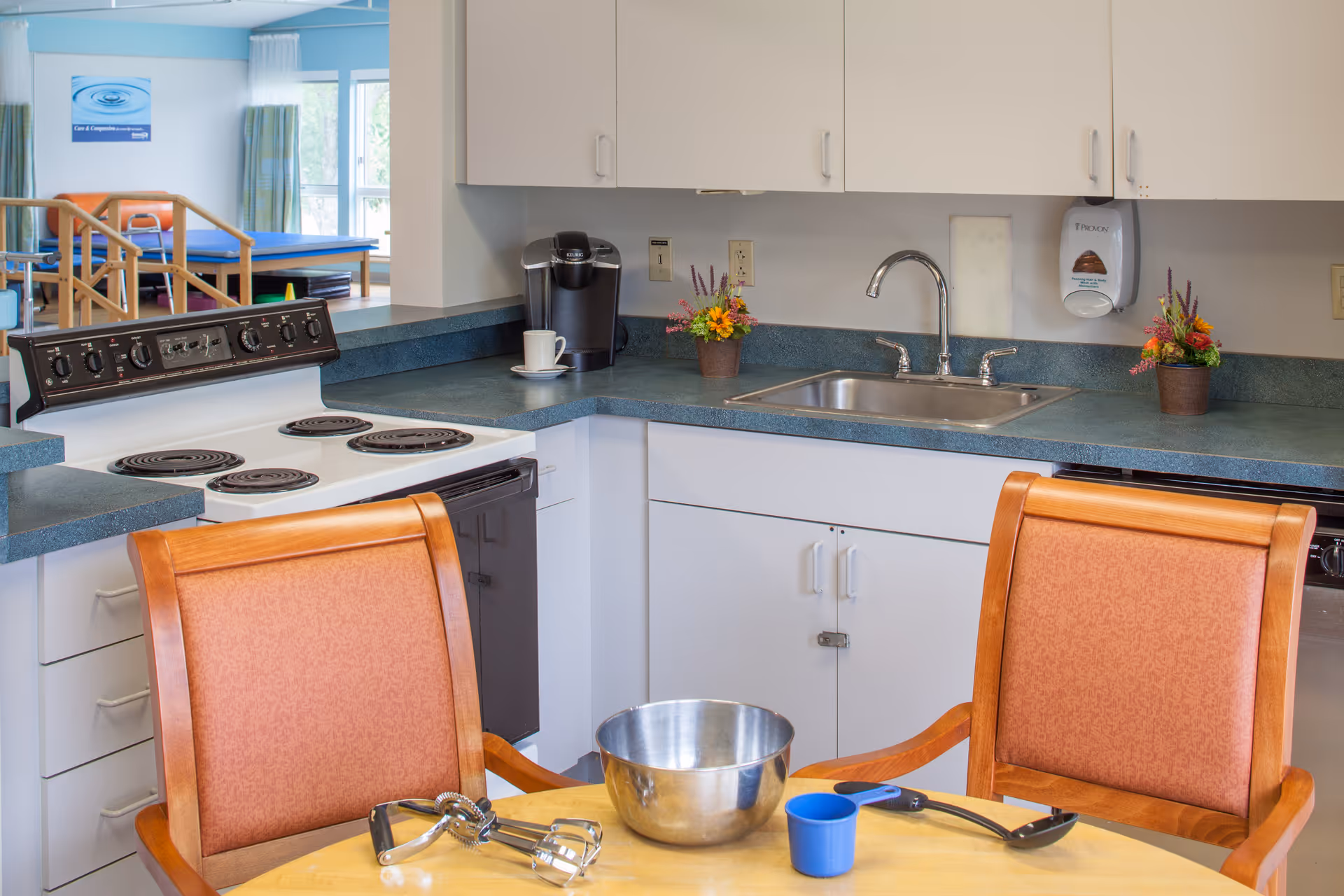 Communal kitchen area with a stove, sink, countertop, and a table with chairs and cooking utensils.