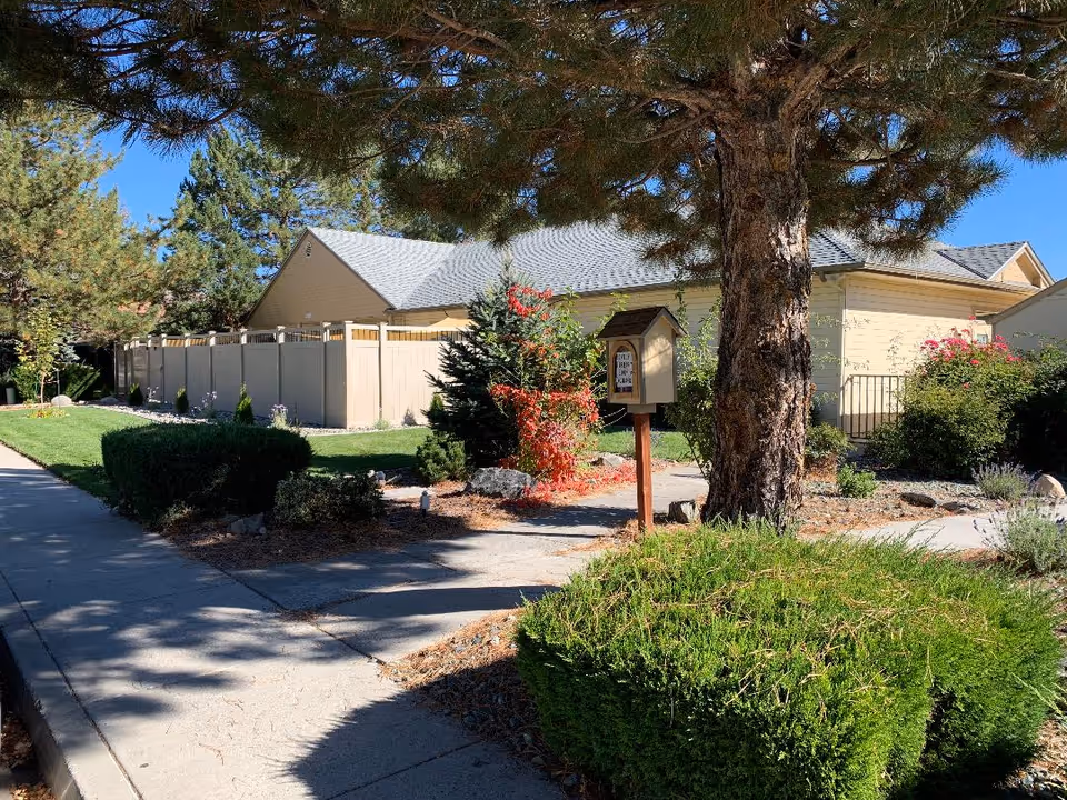 Exterior view of a senior living facility with a beige building, fence, trees, and walkway.