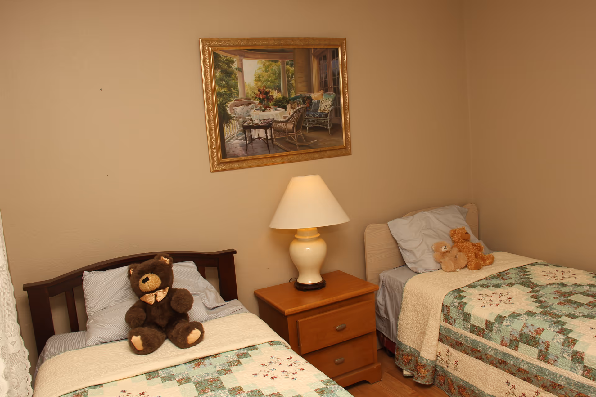 A simple bedroom with two twin beds featuring teddy bears, a nightstand with a lamp, and a framed painting on the wall.