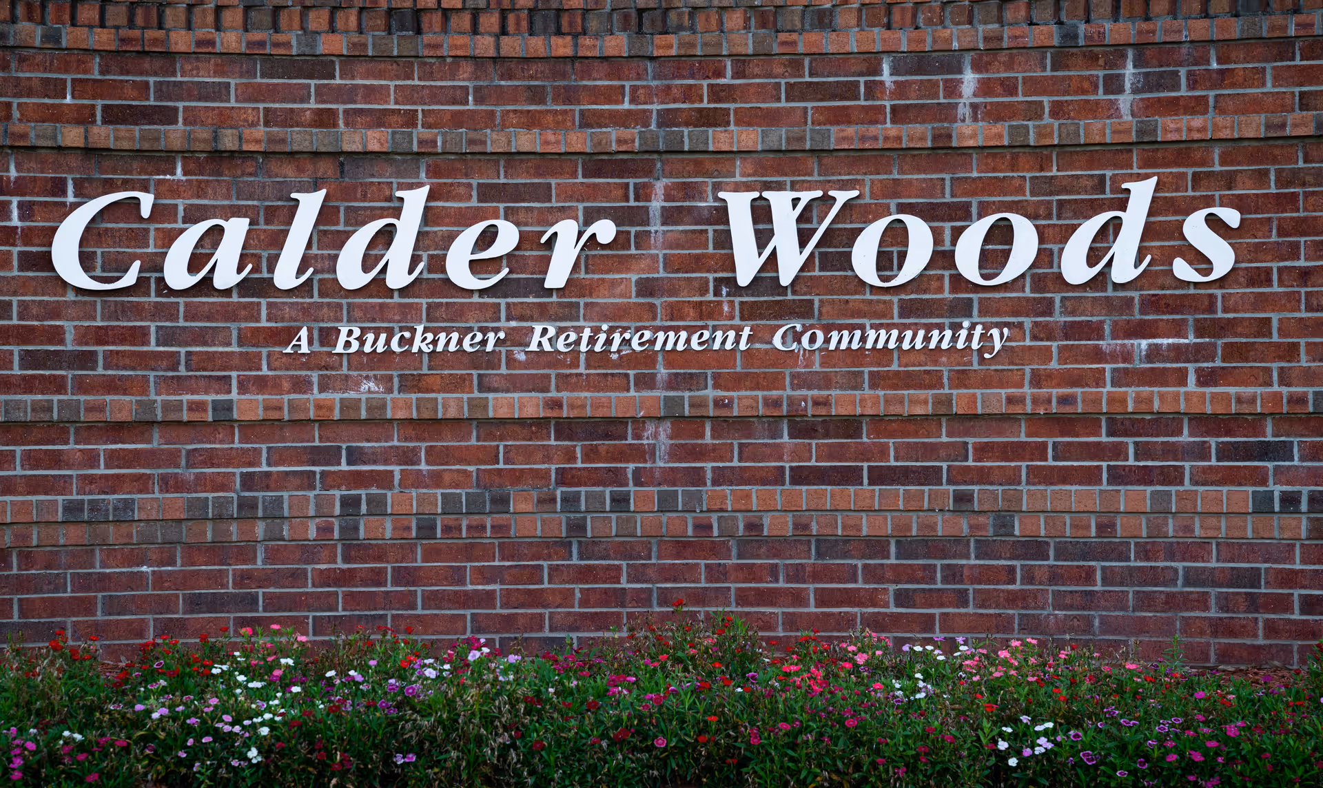 A brick wall with white letters spelling out 'Calder Woods' and below it 'A Buckner Retirement Community', with a flower bed of colorful flowers in front of the wall.