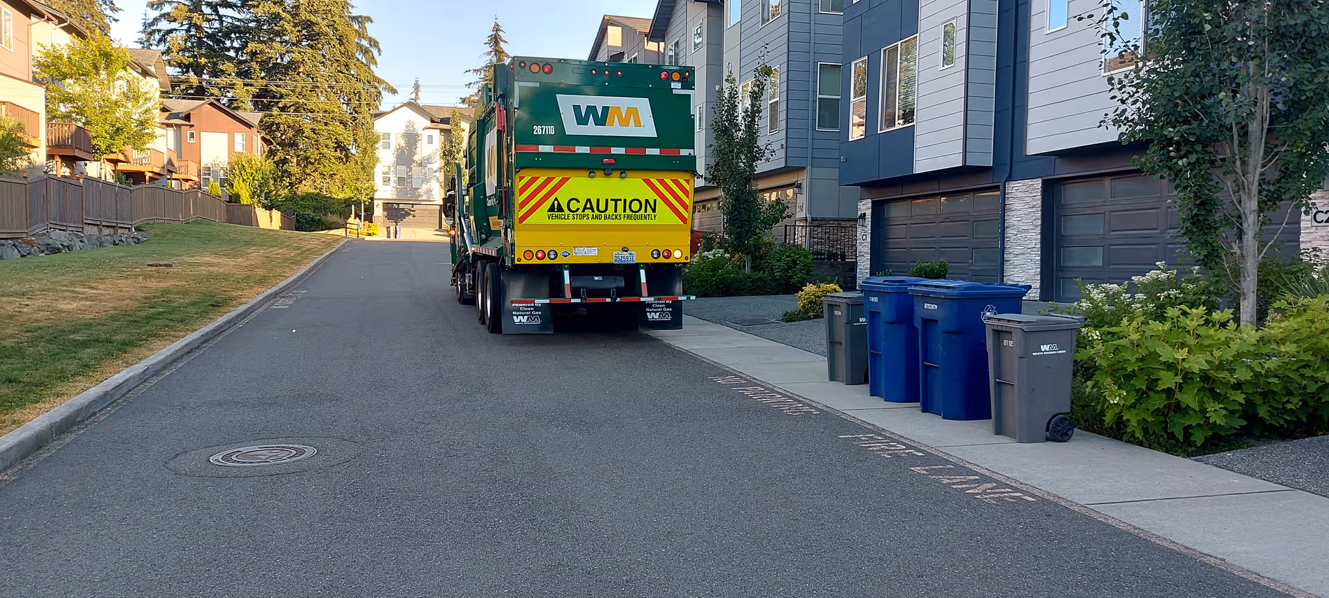 Residential street with a large waste truck beside modern buildings and blue recycling bins.