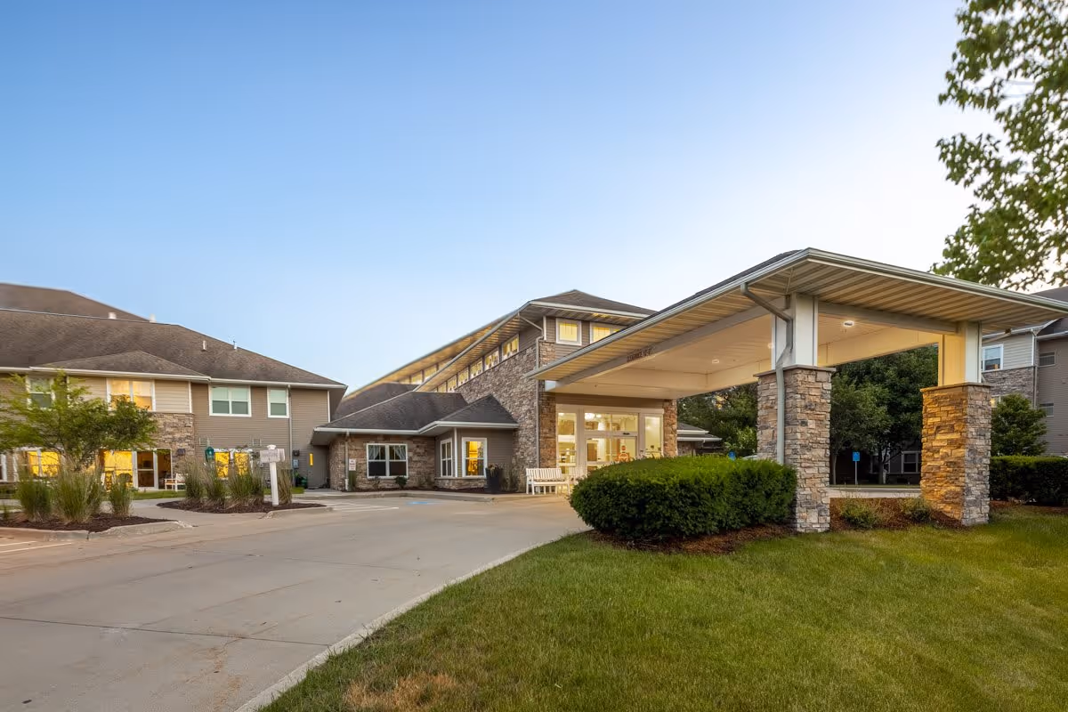 Exterior view of Independence Village of Waukee senior living facility during early evening, showing a covered entrance with stone pillars, well-maintained landscaping, and a multi-story building with lit windows.