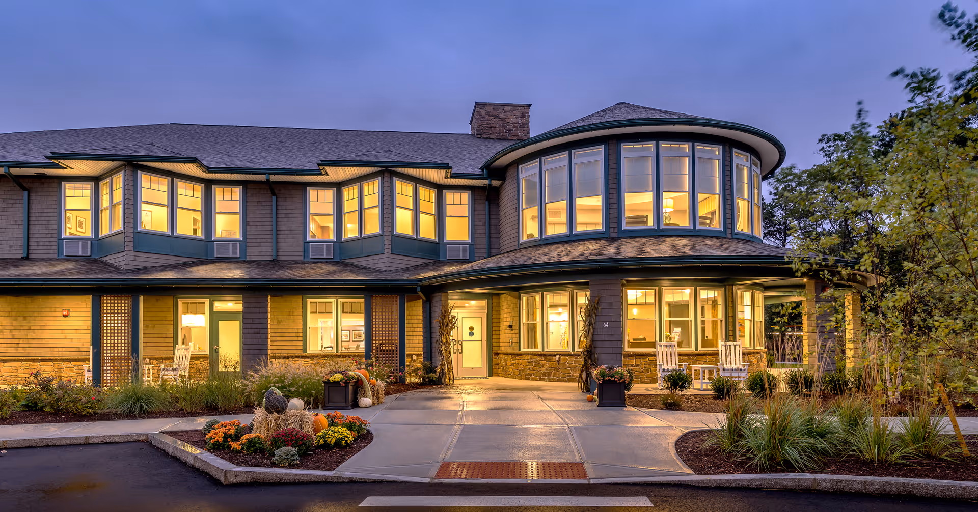 Front entrance of a two-story senior living building lit from within, featuring a round turret of windows, porch rocking chairs, and a landscaped driveway at dusk.