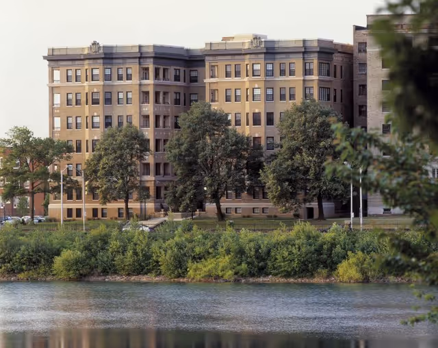 A multi-story brick residential building seen across a river with trees and shrubbery along the shoreline.
