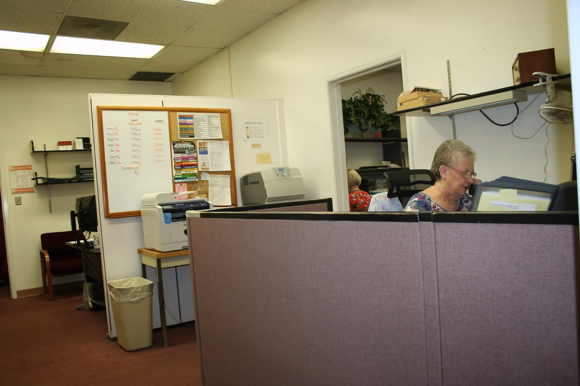 Office reception area with cubicles, a printer, and staff at computer desks.