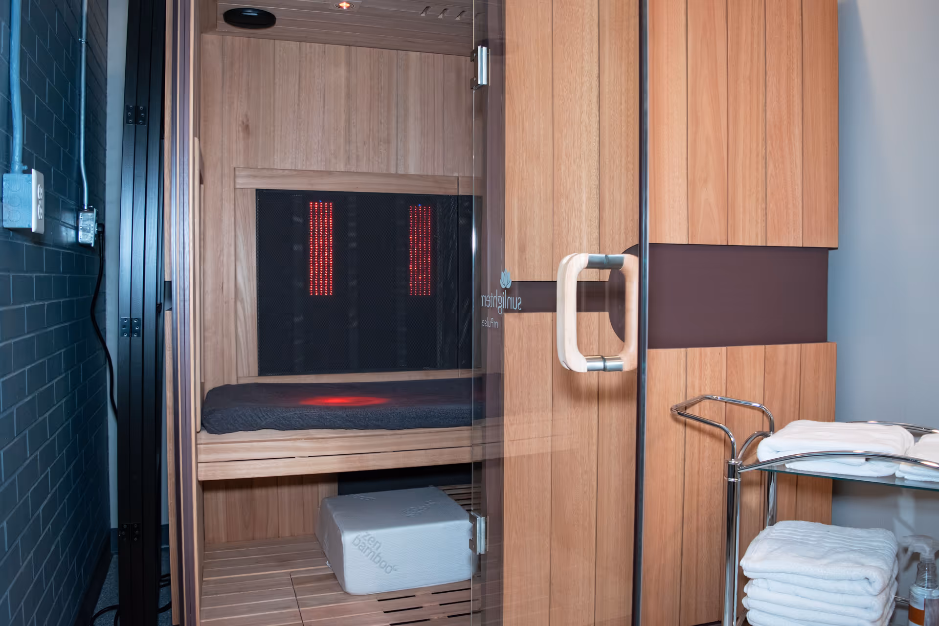 Wooden infrared sauna inside a facility, glass door, bench, and towel cart nearby.
