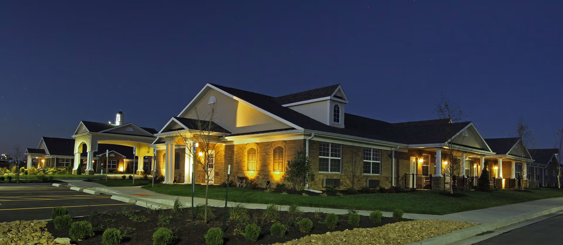 Exterior view of Benton House of Raymore senior living facility at night, showing a well-lit brick building with white columns, a covered entrance, landscaped greenery, and an empty parking lot.
