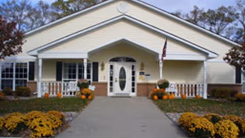 Front exterior view of a single-story building with a white door and windows, a small porch with railings, and landscaping featuring yellow flowers and pumpkins on either side of the walkway leading to the entrance.