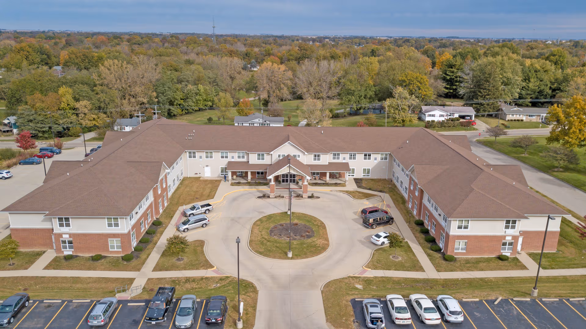 Aerial view of a large, U-shaped senior living facility building with a brown roof and brick and beige exterior walls. The building is surrounded by a parking lot with several cars parked, and there is a circular driveway with a small grassy island in the center. The background shows a wooded area with trees displaying fall colors under a clear sky.