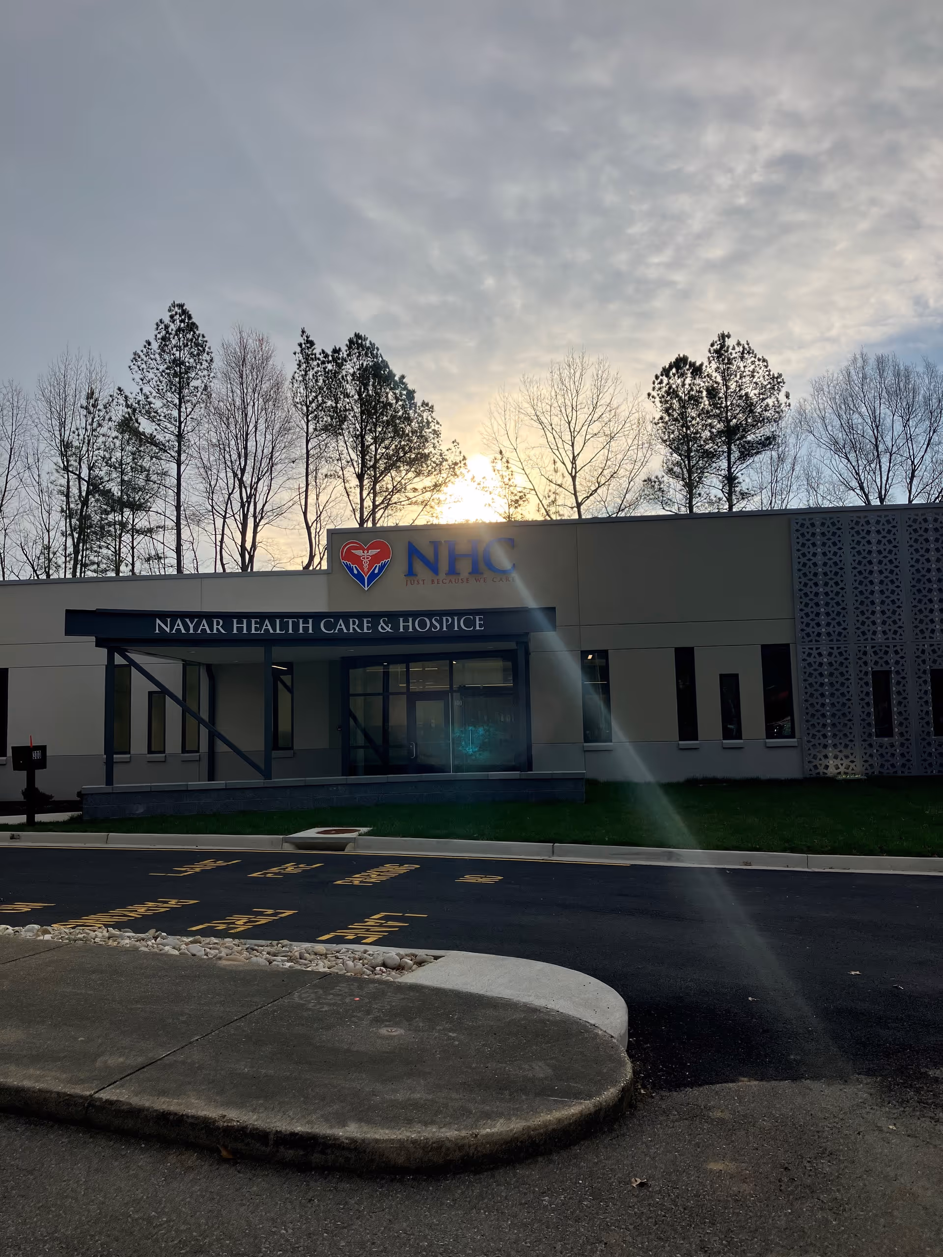 Front view of Nayar Health Care & Hospice building with sign and sun behind trees.