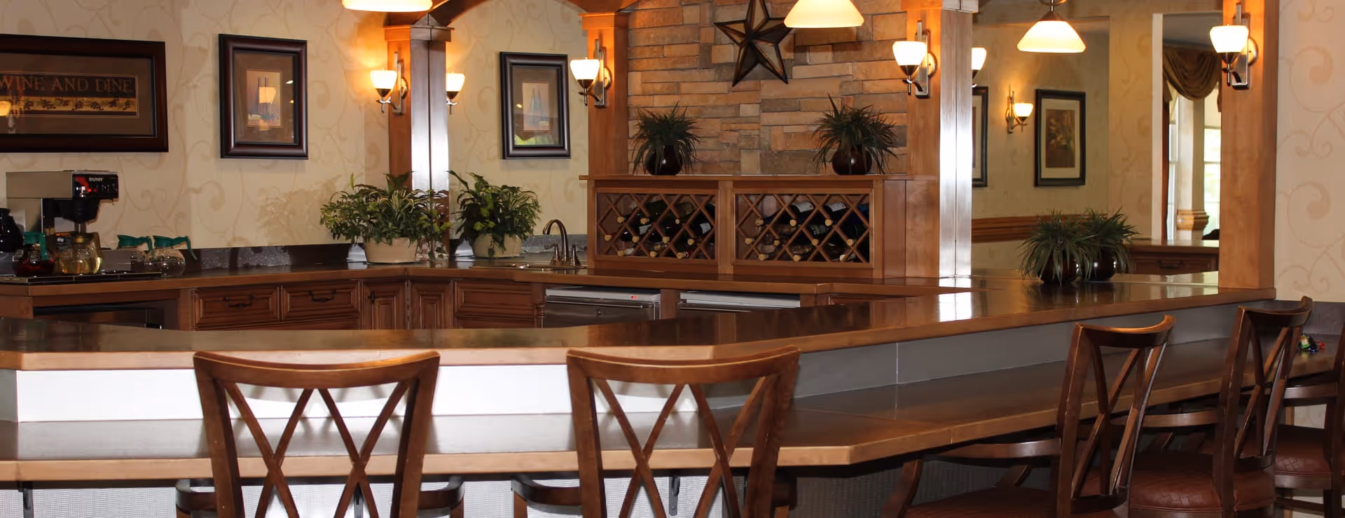 Interior view of a dining area with a long wooden counter and several wooden chairs with cross-back designs. Behind the counter is a kitchen area with a wine rack, plants, framed pictures on the wall, and warm lighting fixtures including wall sconces and hanging pendant lights. The decor features wood accents and a stone wall with a star decoration.