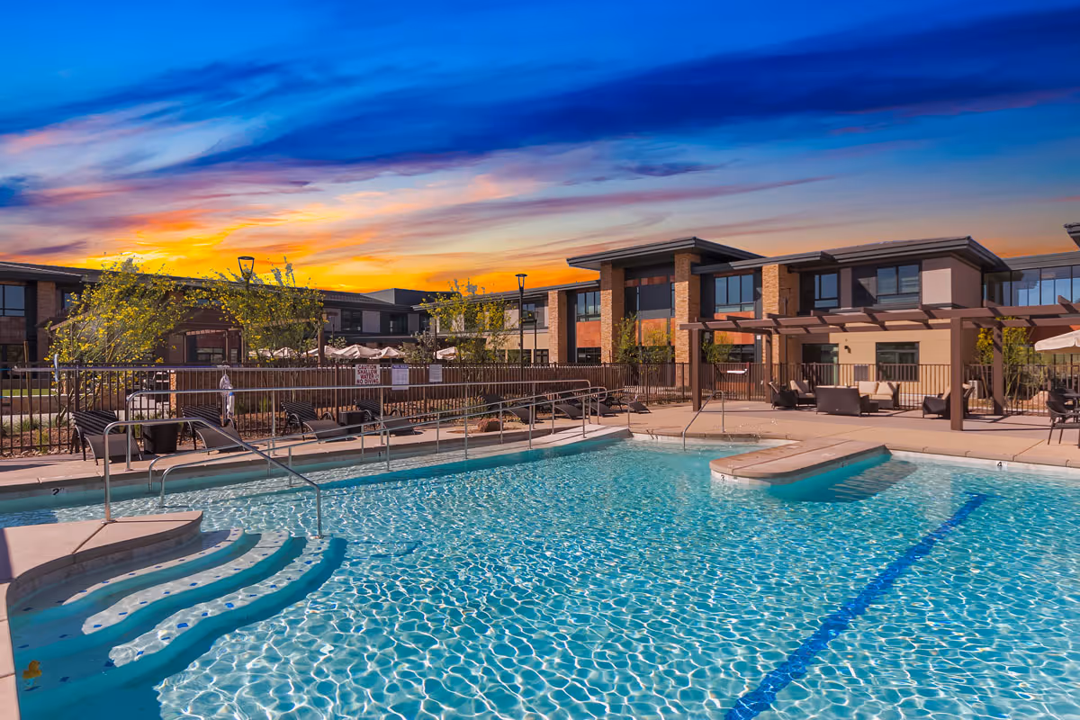 Outdoor swimming pool with clear blue water and steps leading into the pool, surrounded by lounge chairs and tables with umbrellas. Modern two-story buildings and trees are visible in the background under a colorful sunset sky.