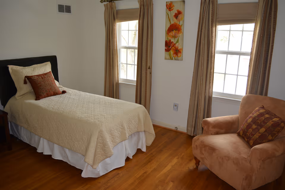 A tidy bedroom with a bed, armchair, two windows, curtains, and wooden flooring.