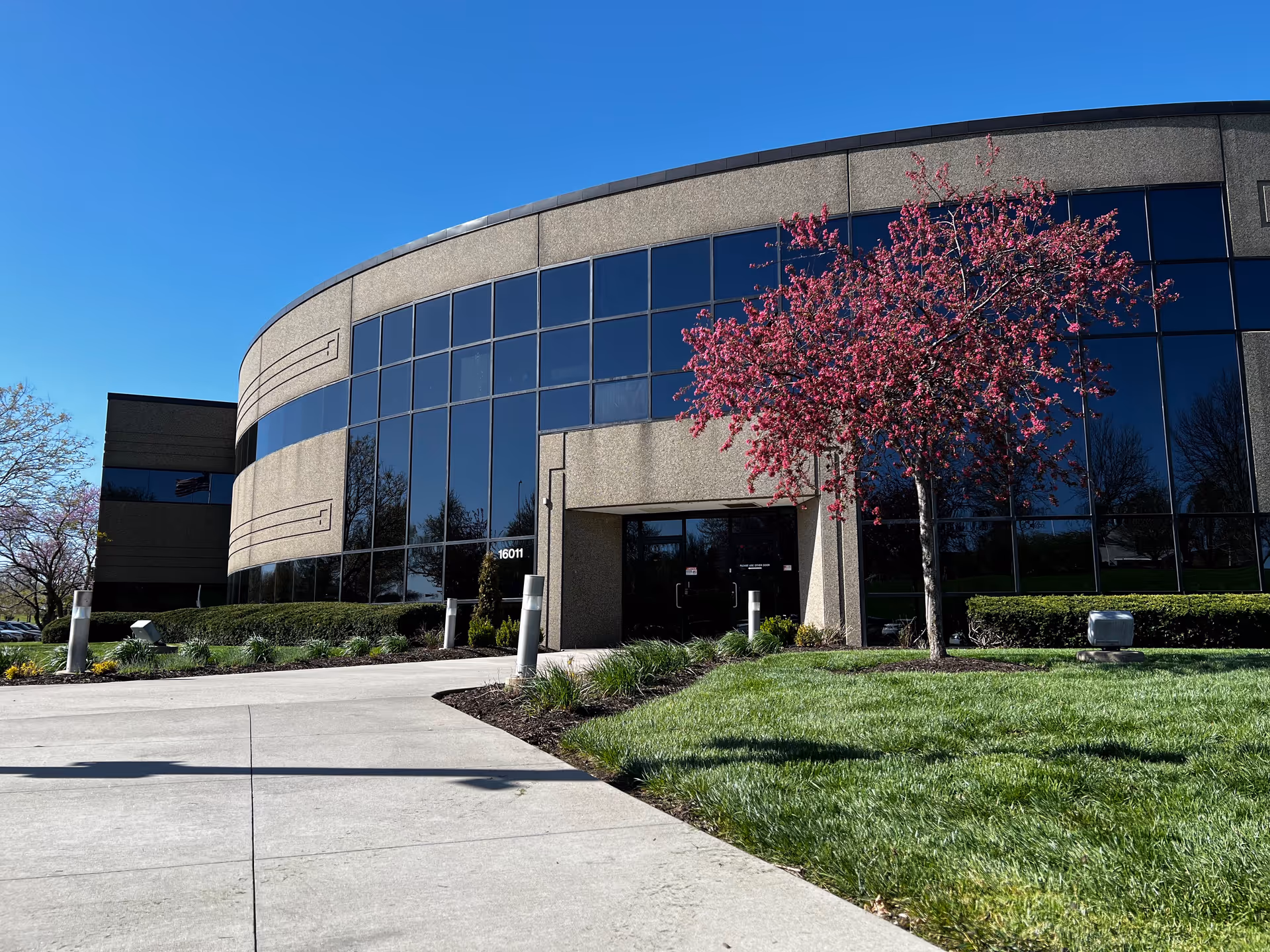 Curved glass-front building with entryway, landscaping, and blooming pink tree