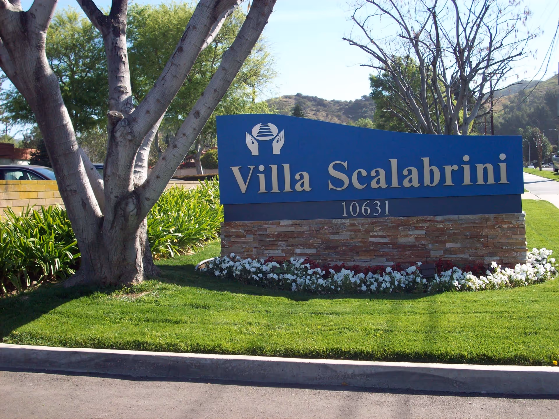 Entrance sign reading "Villa Scalabrini 10631" on a blue marker surrounded by grass, flowers, and trees.