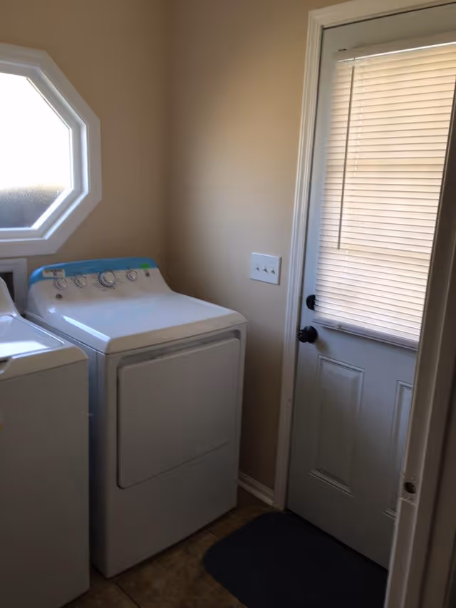 Laundry room with a white washing machine and dryer side by side, a small octagonal window on the left wall, and a door with closed blinds on the right.