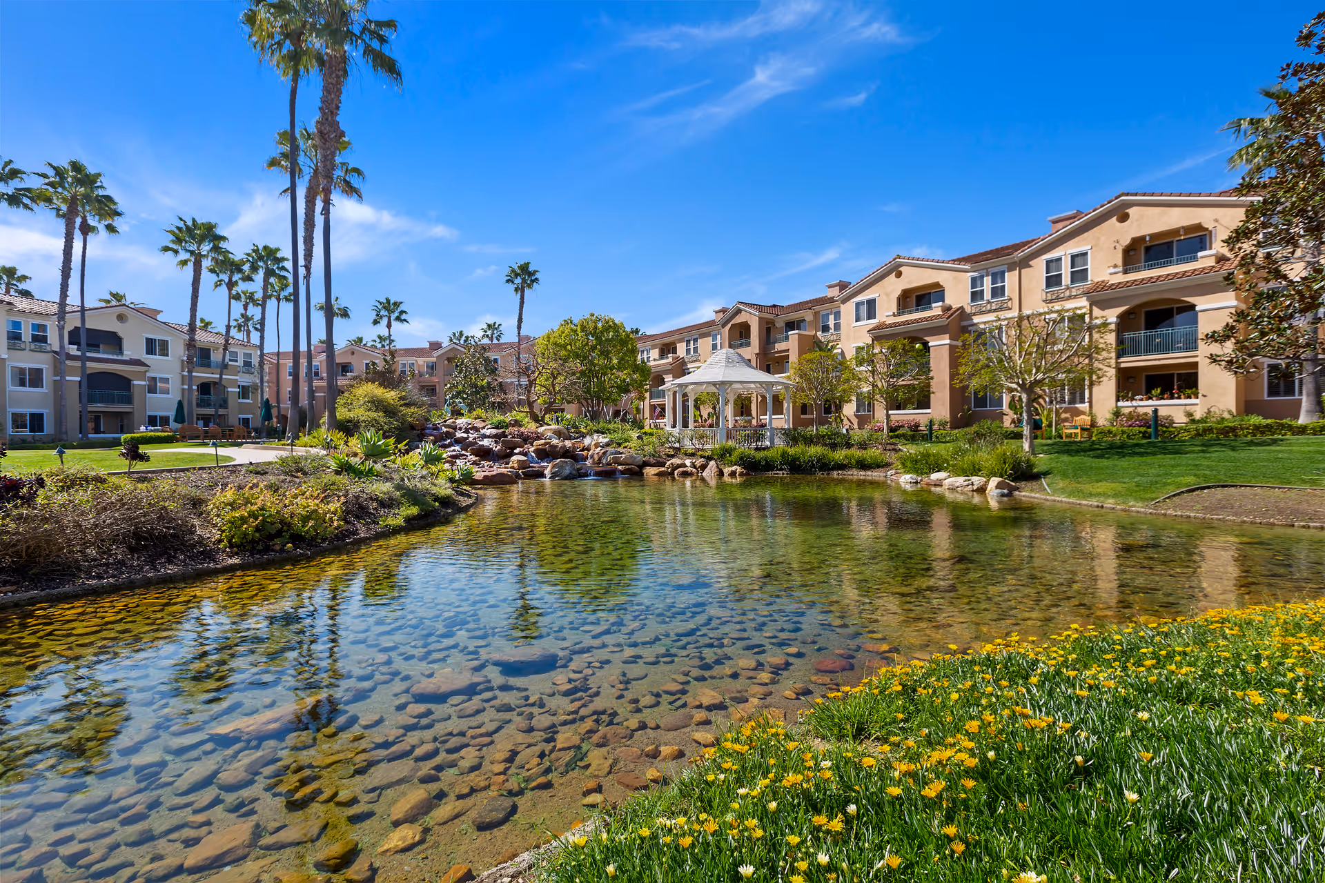 Landscaped pond with clear water, yellow flowers, palm trees, and a gazebo in front of a multi-story senior living complex under a blue sky.