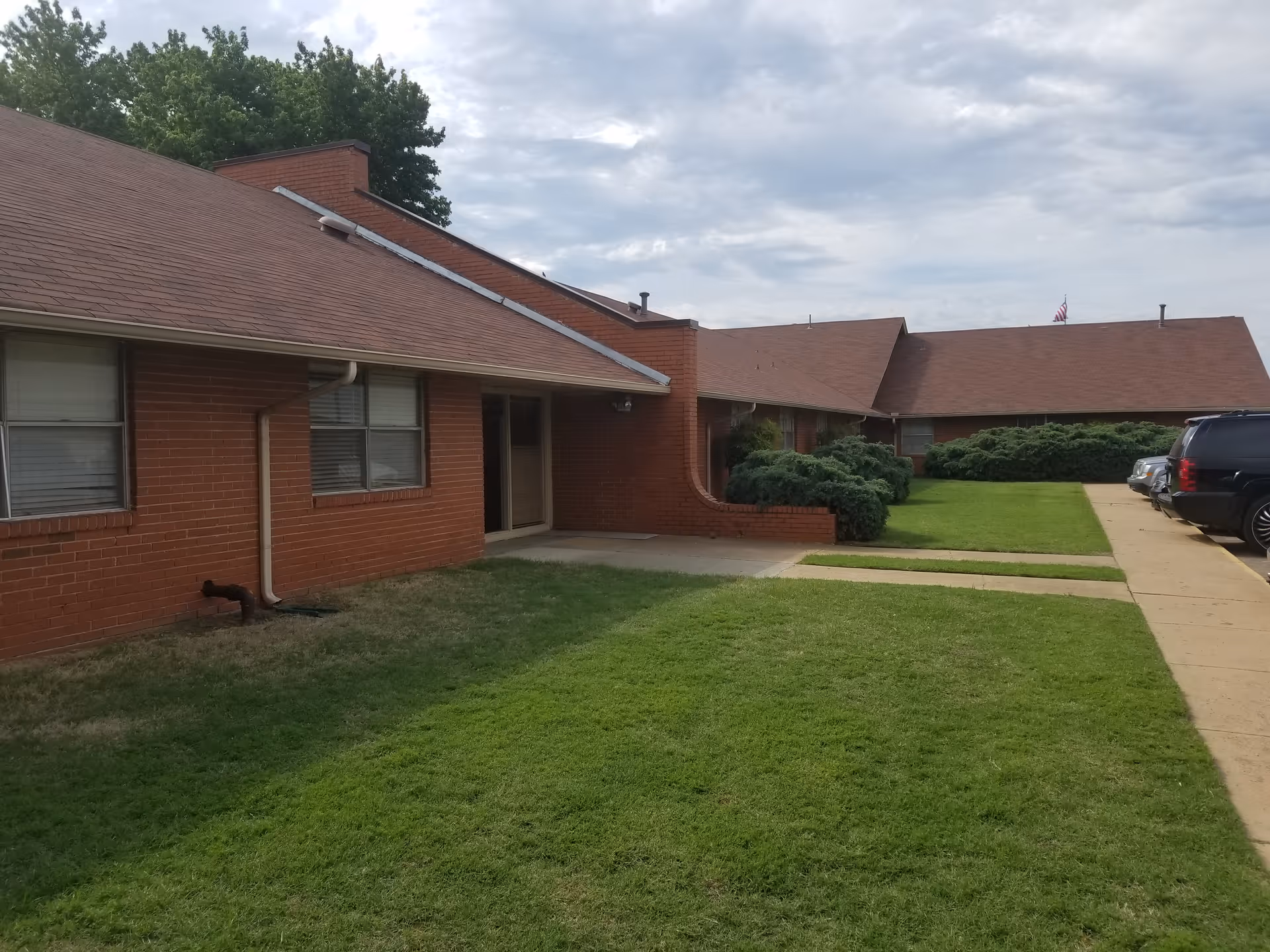 Exterior view of a single-story brick building with a sloped roof, green lawn, bushes, and a sidewalk leading to the entrance. Several parked cars are visible on the right side under a cloudy sky.