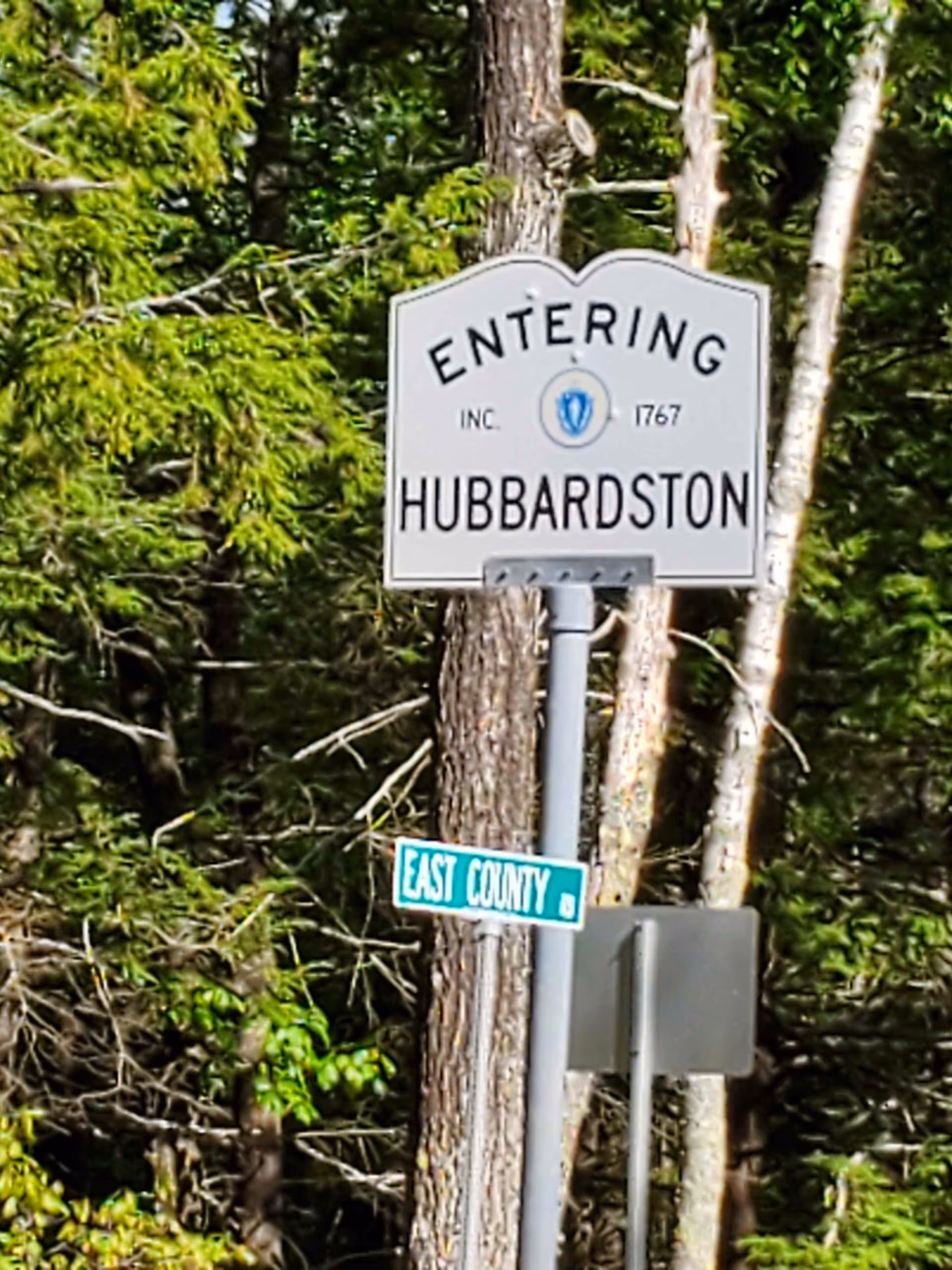 A highway sign reading Entering Hubbardston among trees, with a green East County street sign nearby.