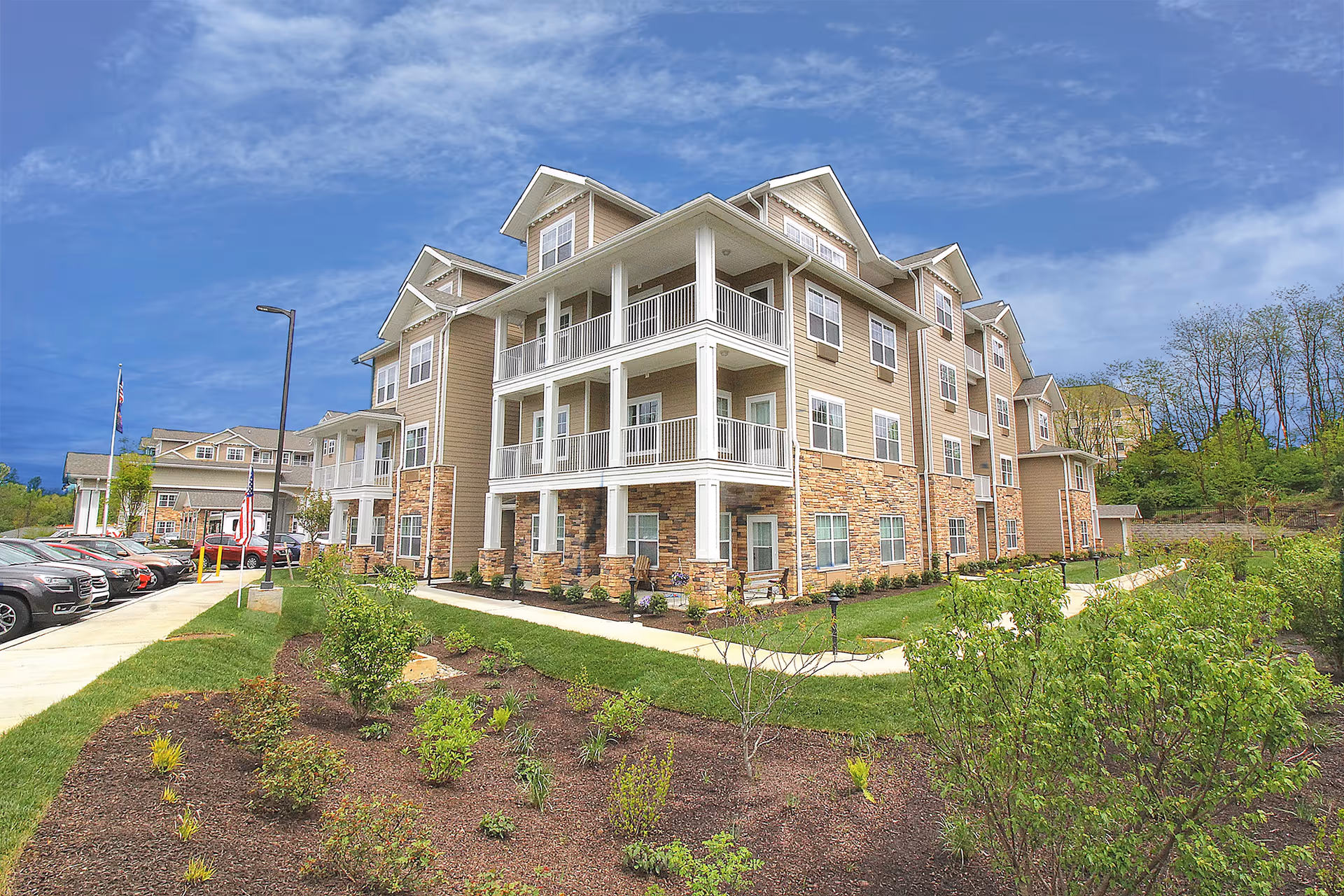 Three-story beige senior living building with balconies, landscaped grounds, and parked cars under a blue sky.