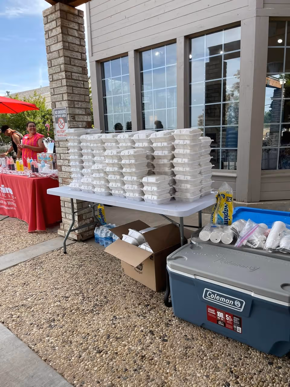 Outdoor table with stacked foam food containers beside a cooler near a building.