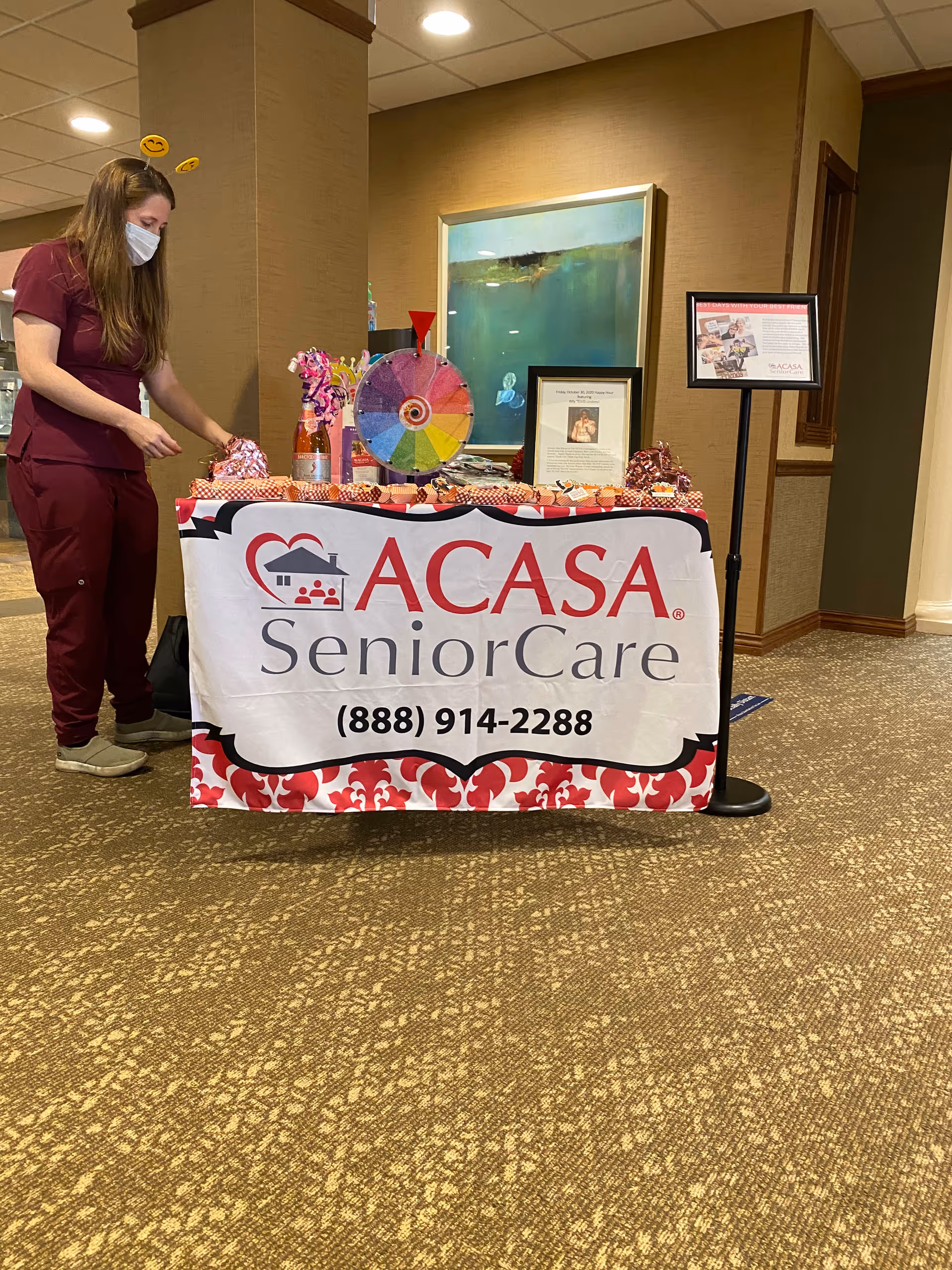 A caregiver in scrubs and mask stands beside an ACASA SeniorCare table display indoors.