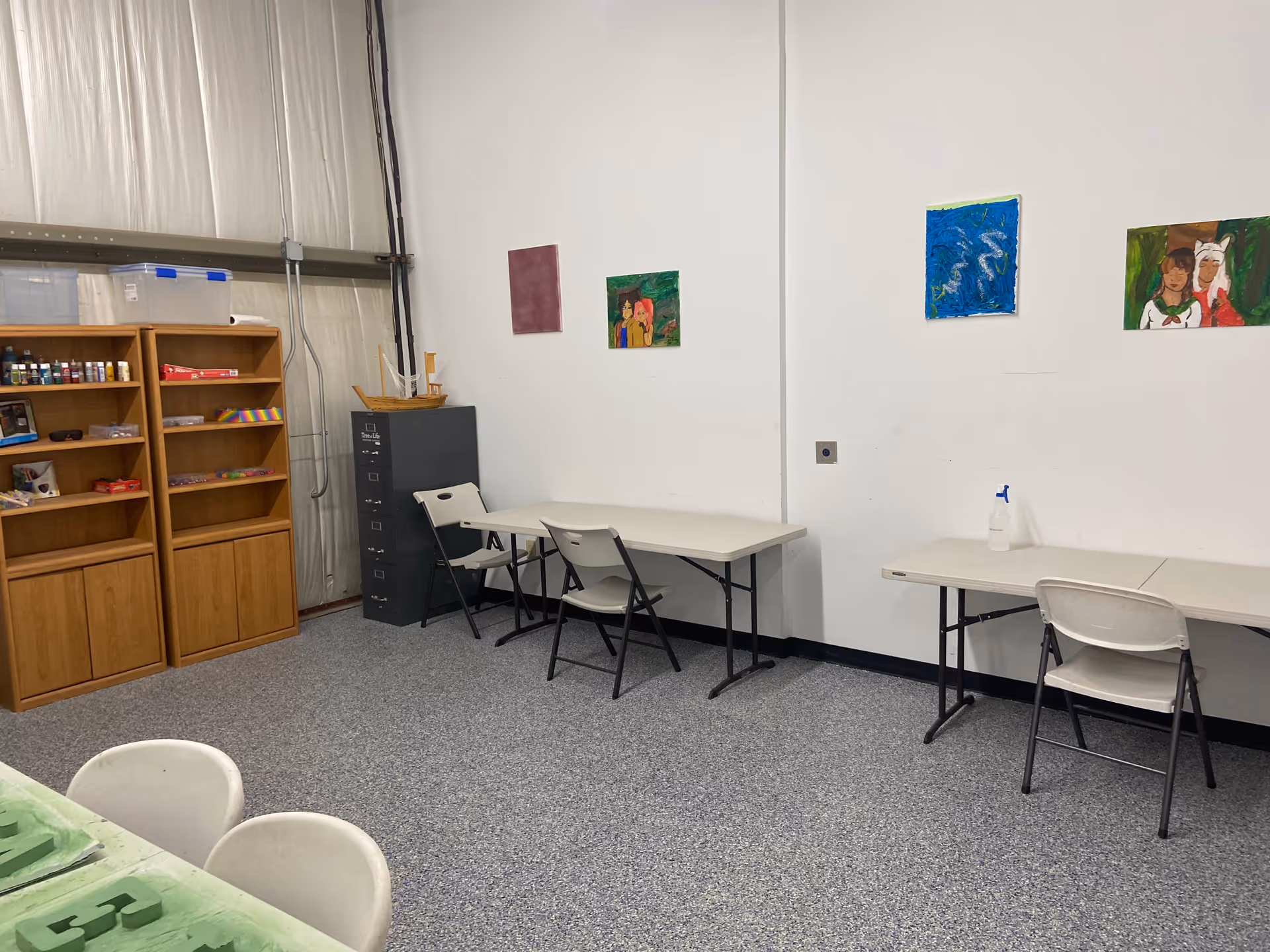 A small craft room with tables, chairs, shelves, and art supplies along a white wall.