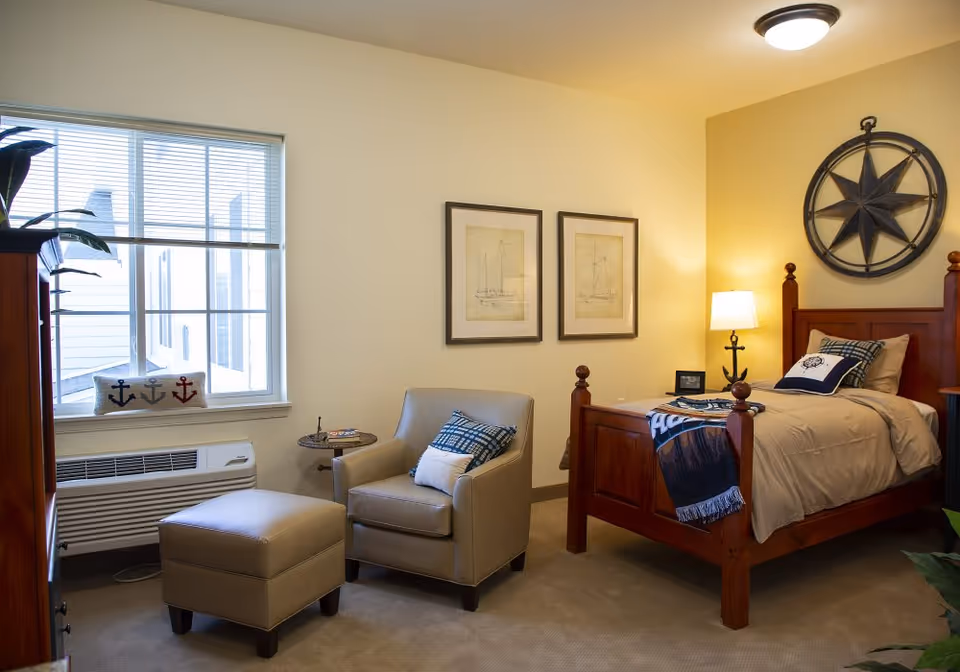 A cozy, well-lit bedroom featuring a wooden single bed with nautical decor, an armchair and ottoman, a window, and a bedside lamp.