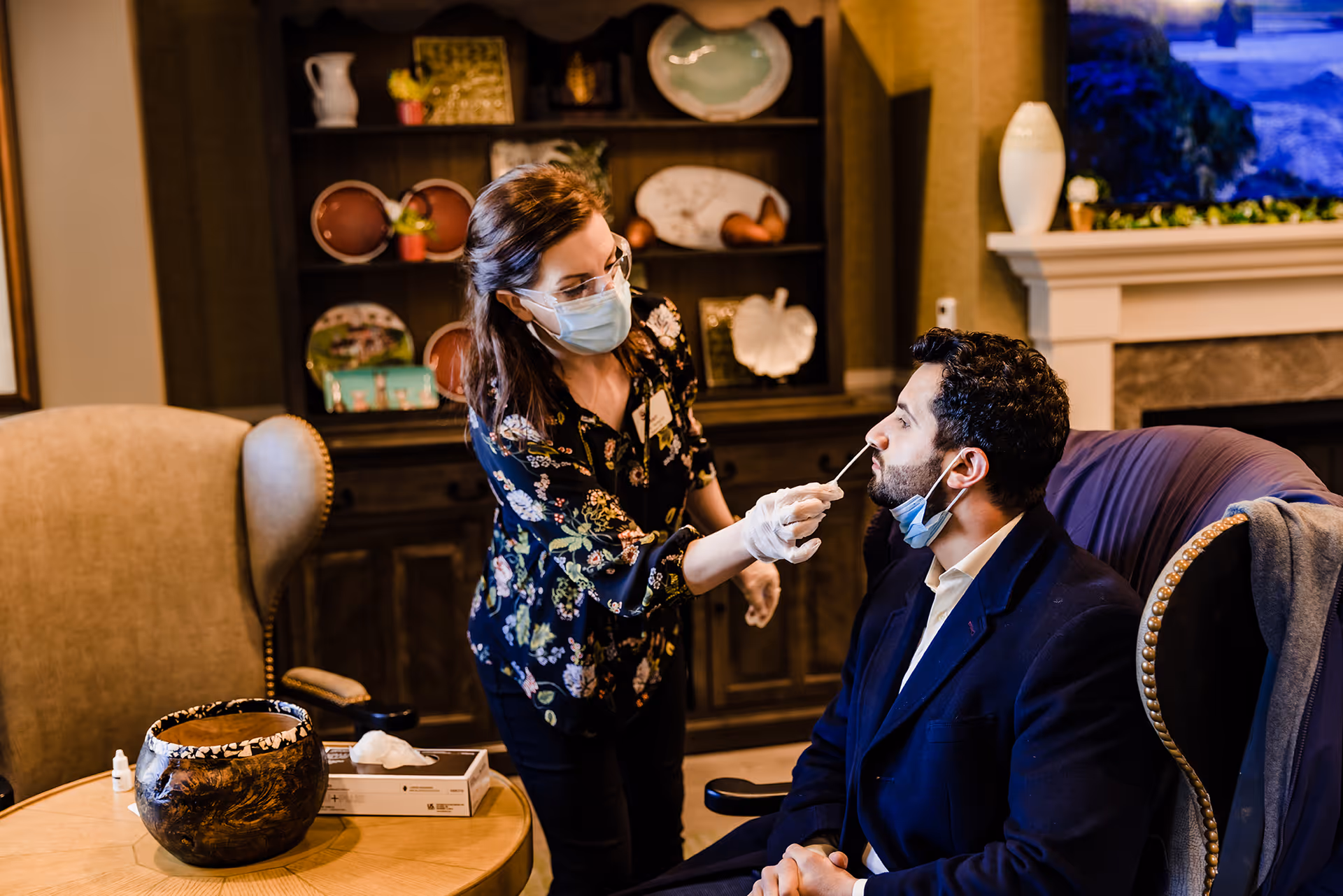 A woman wearing a face mask and gloves is administering a nasal swab test to a seated man in a cozy room with a fireplace and decorative shelves in the background.