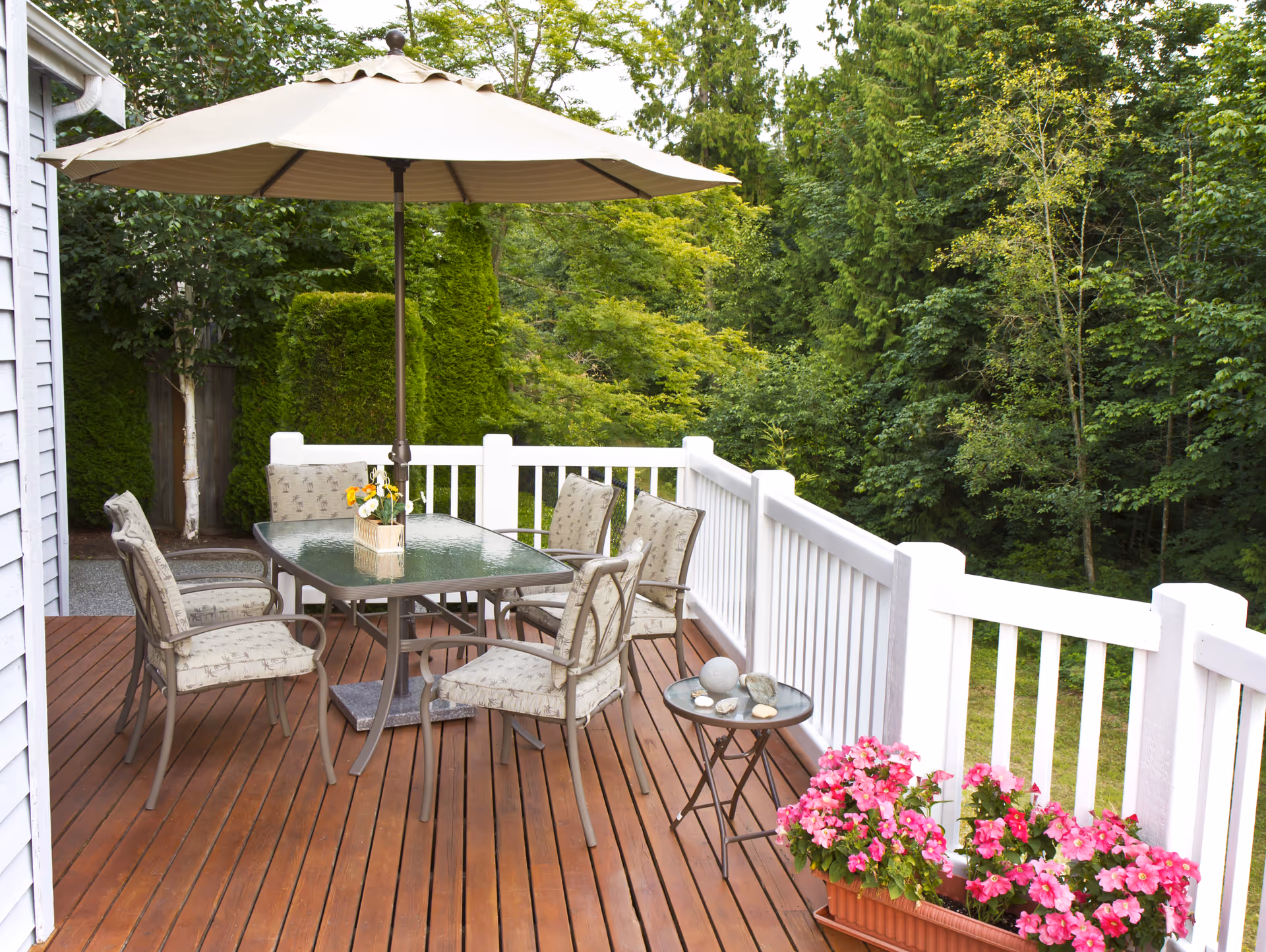 Outdoor wooden deck with glass-table dining set, umbrella, and flowering planters.