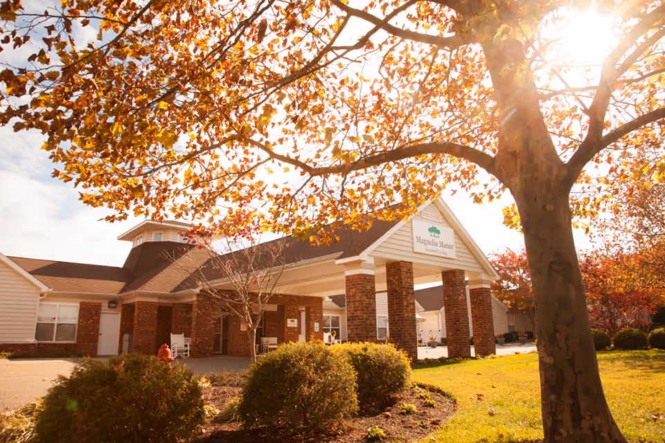 Exterior view of a senior living facility named Magnolia Manor Assisted Living with a brick entrance and white pillars, surrounded by bushes and trees with autumn leaves under a sunny sky.