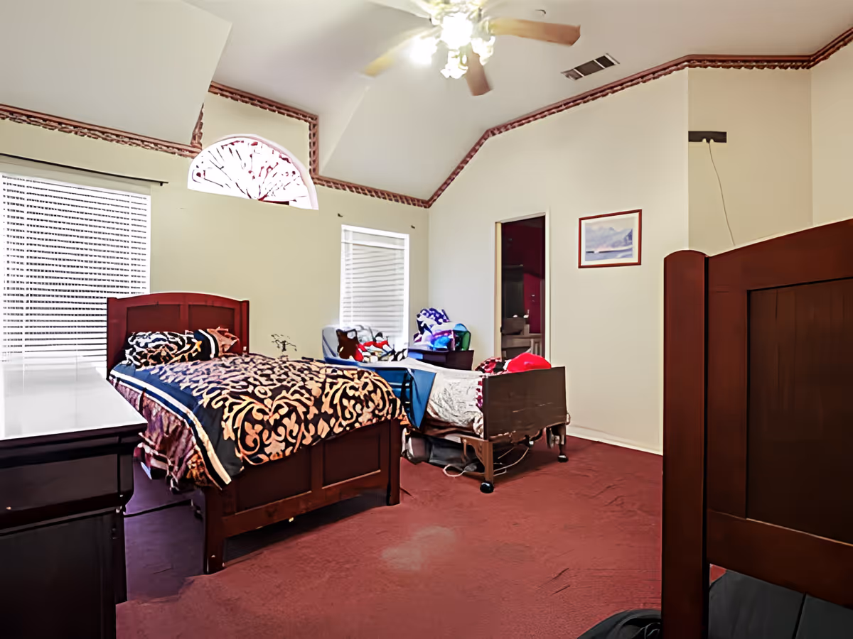 A bedroom with two beds, one wooden bed with a patterned blanket and another hospital-style bed with blankets and pillows. The room has a maroon carpet, cream-colored walls with decorative trim near the ceiling, two windows with blinds, a ceiling fan with lights, and a framed picture on the wall. There is also a chair with stuffed animals and blankets near the window.