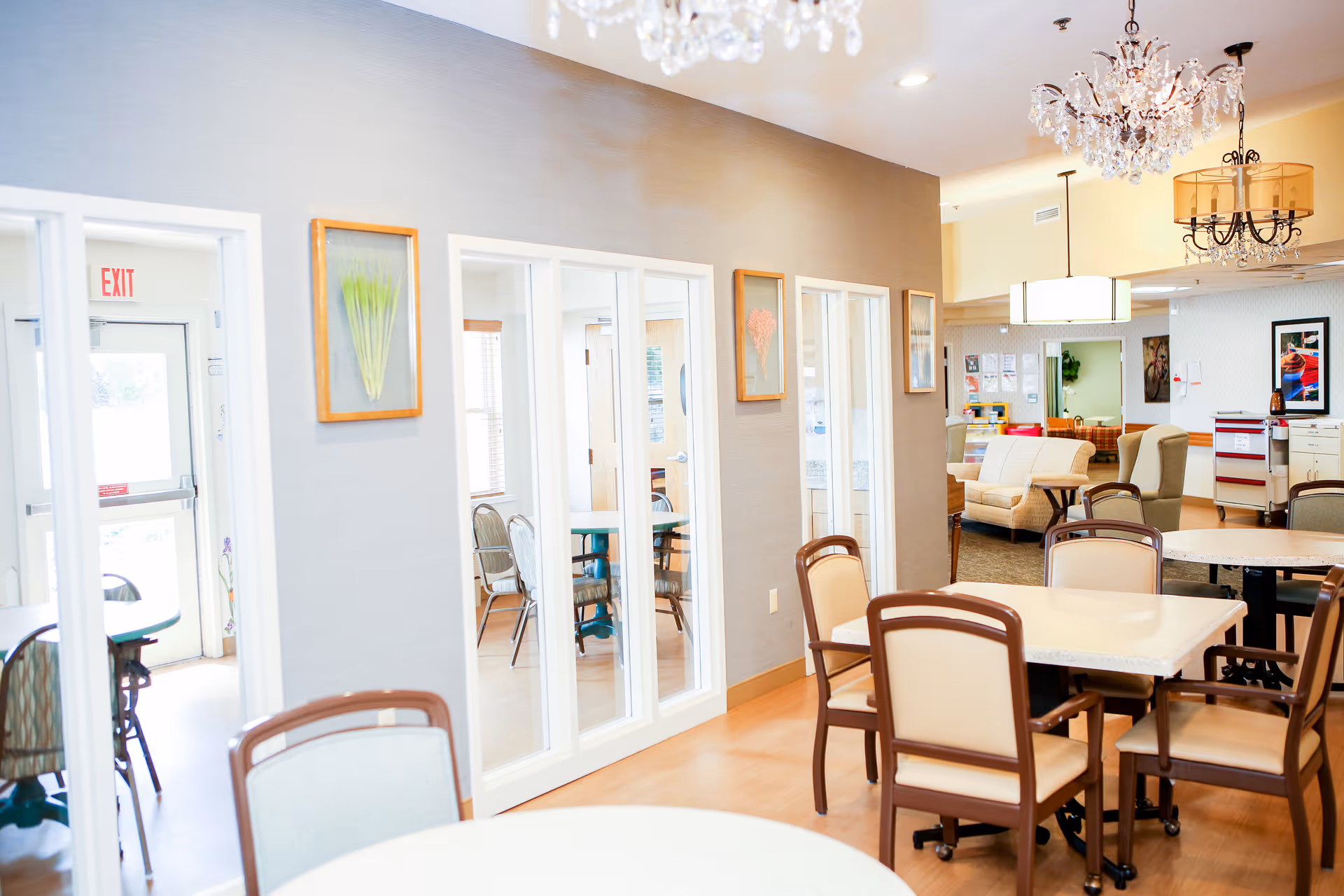 Bright senior living dining area with round tables, beige chairs, and glass-door partitions.