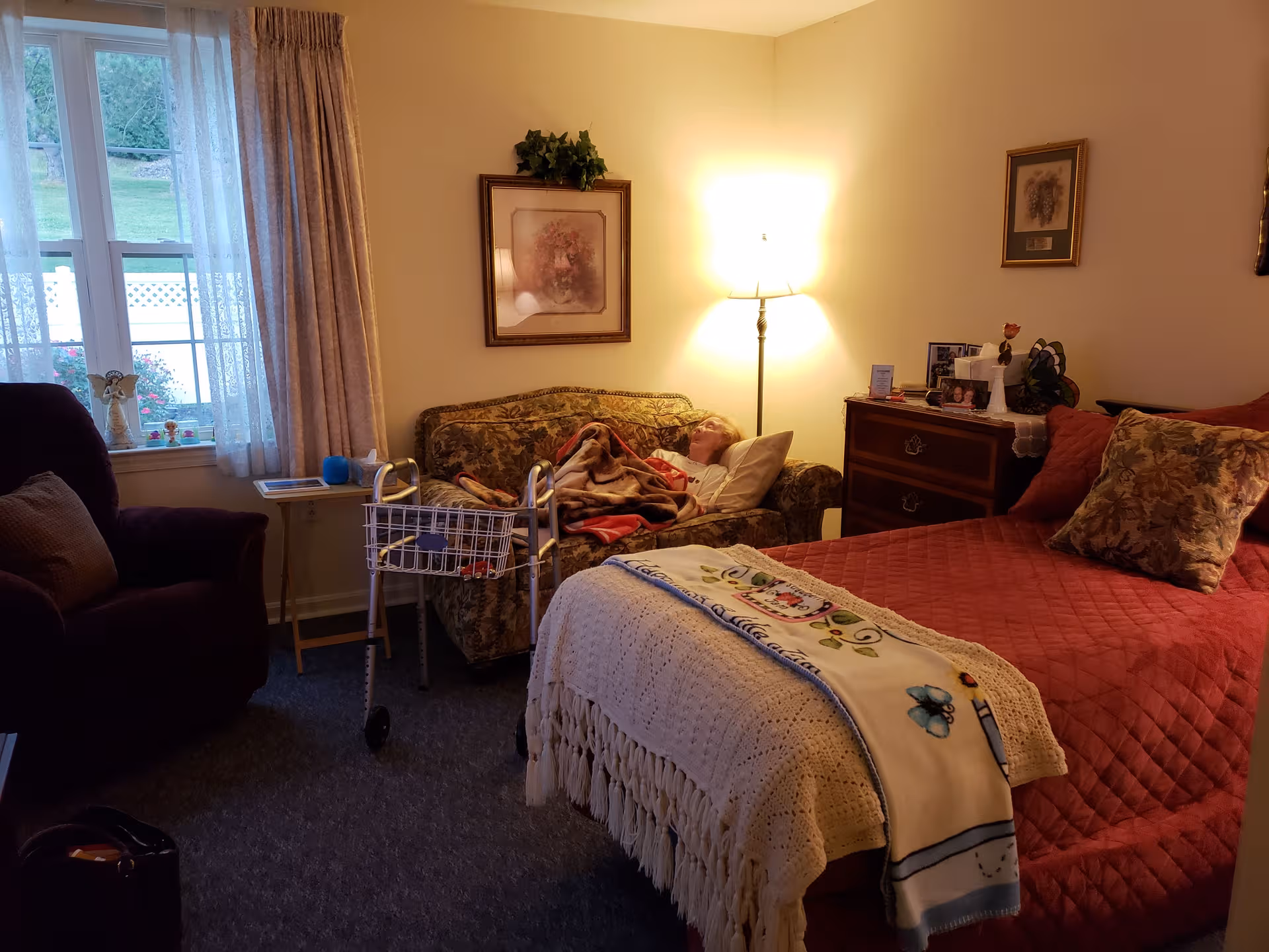 Senior living bedroom with bed, floral sofa, walker, nightstand, and warm lamp.