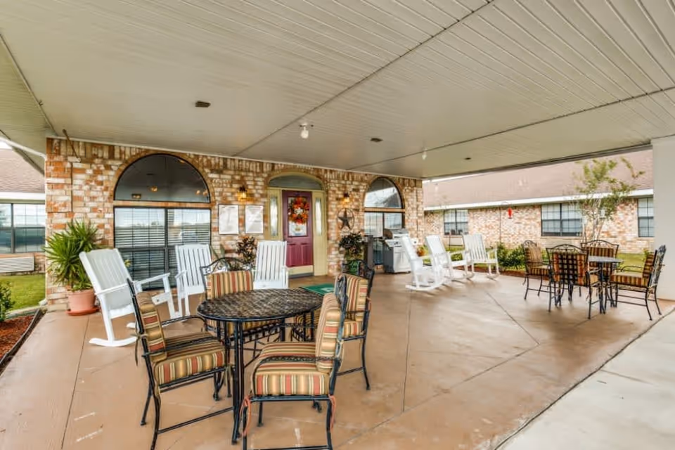 Covered front entrance patio of a senior living facility with multiple outdoor tables, striped cushioned chairs, white rocking chairs, and a brick façade with a maroon door.