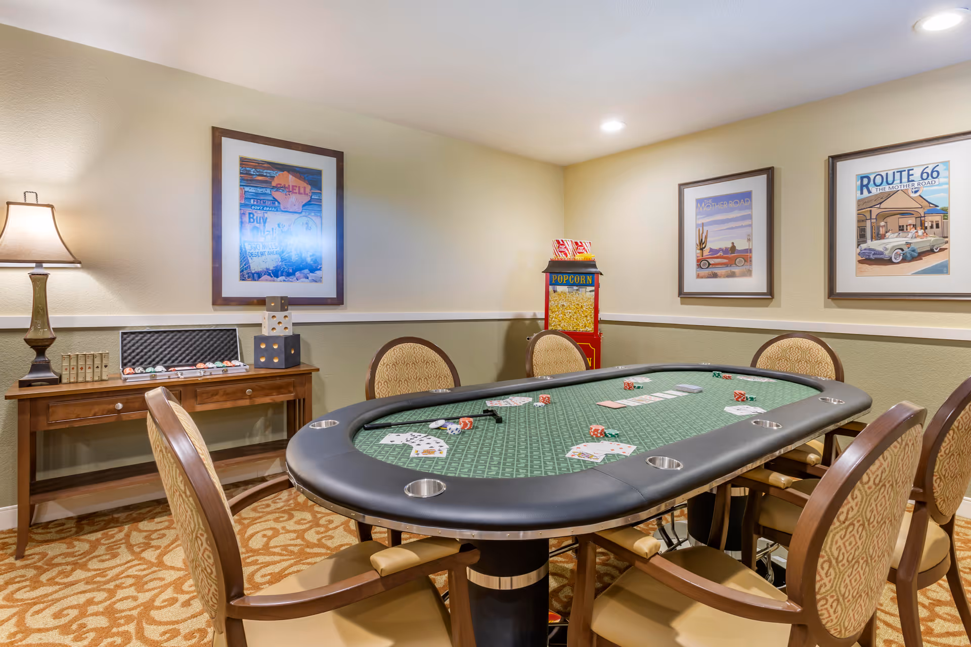 A game room with a green poker table surrounded by six chairs. The table has playing cards and dice on it. Behind the table, there is a wooden console table with a lamp, oversized dice, and poker chips. The walls are decorated with framed vintage-style Route 66 and desert road posters. A popcorn machine is placed in the corner of the room.