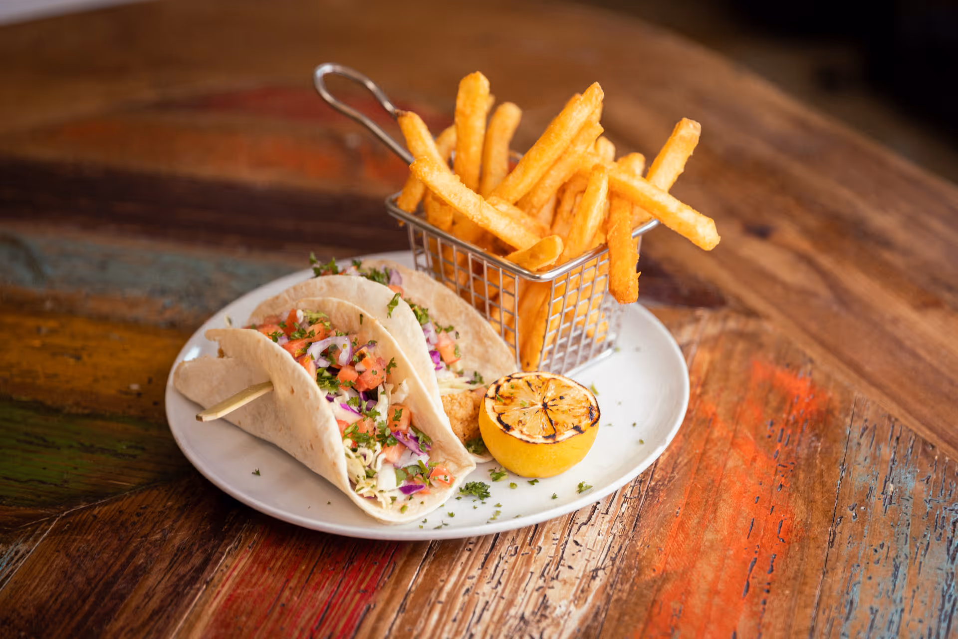 A white plate on a rustic wooden table holding two soft tacos filled with shredded cabbage, diced tomatoes, and herbs, accompanied by a grilled lemon half and a small metal basket filled with French fries.