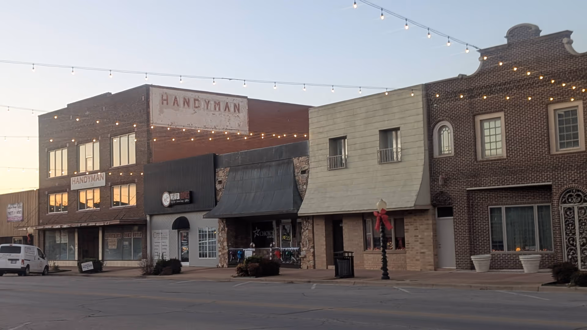 Row of brick storefronts along a street with string lights and a parked van.