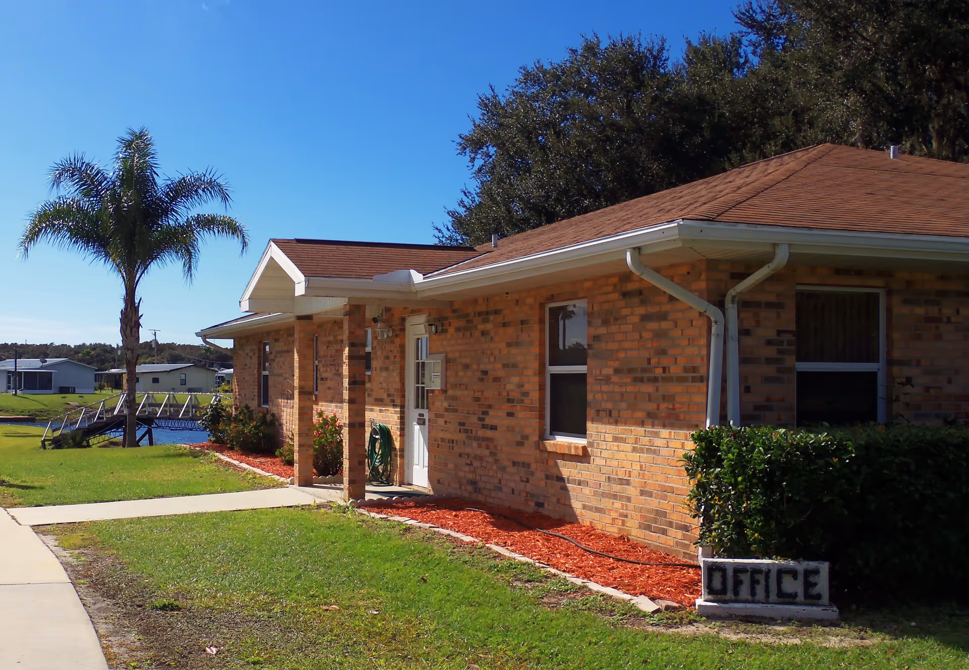 Exterior view of a single-story brick building with a brown roof, a small covered entrance, and a sign that reads 'OFFICE' near a green bush. There is a palm tree and a small bridge over water in the background under a clear blue sky.