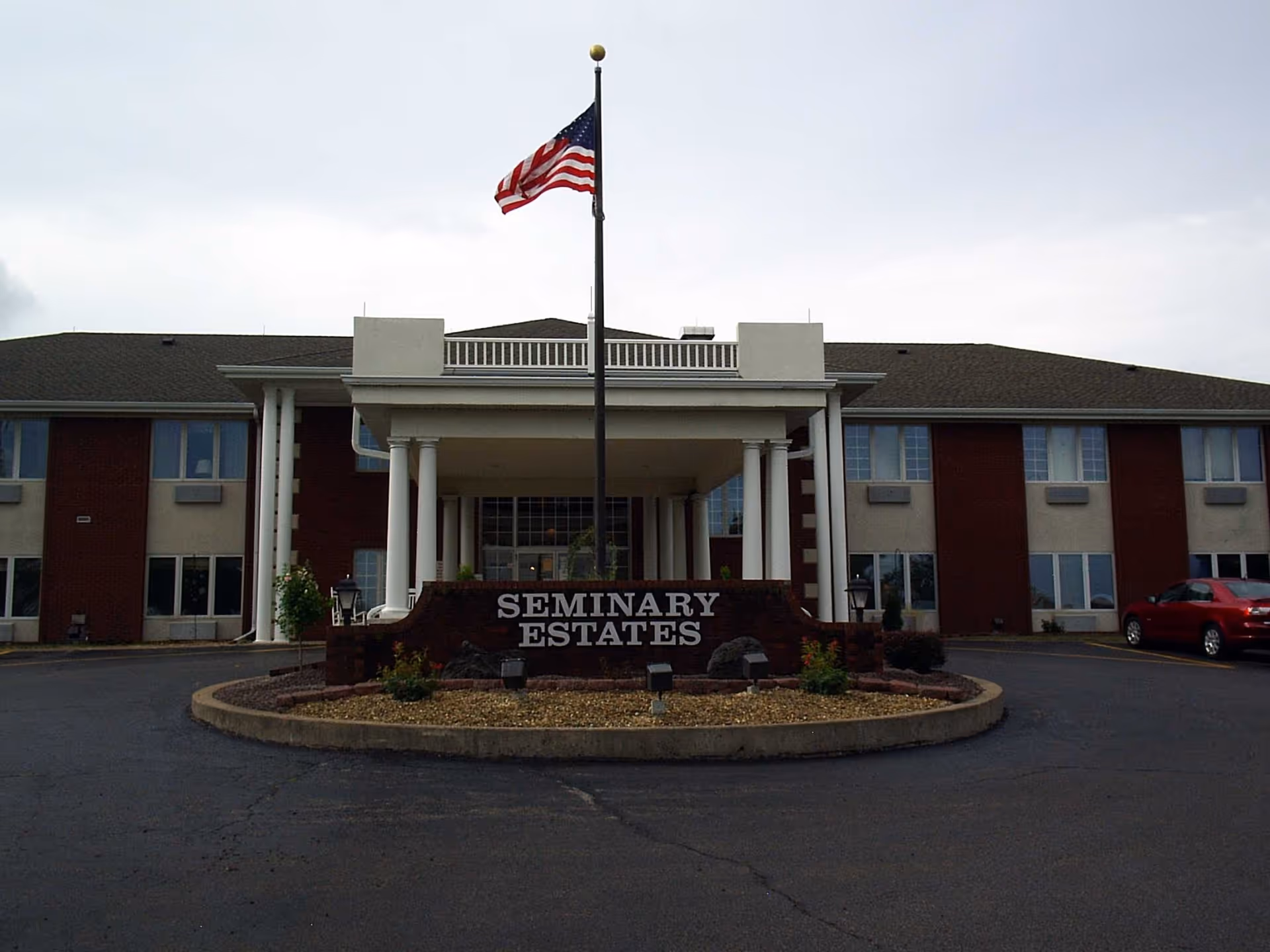 Front exterior view of Seminary Estates building with a covered entrance supported by white columns, an American flag on a flagpole, and a sign reading 'Seminary Estates' in front of the entrance.