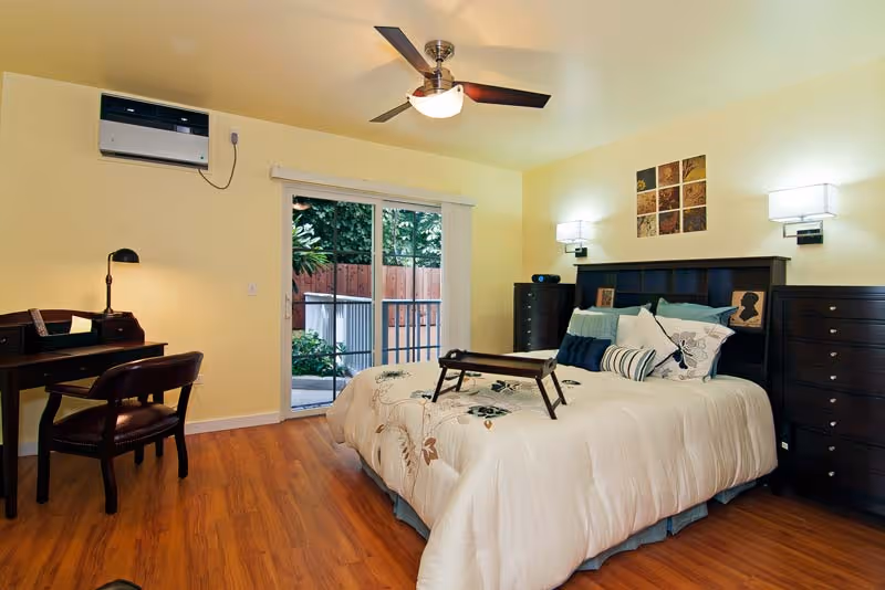 A well-lit bedroom with a large bed, dresser, desk, and sliding door to outside.