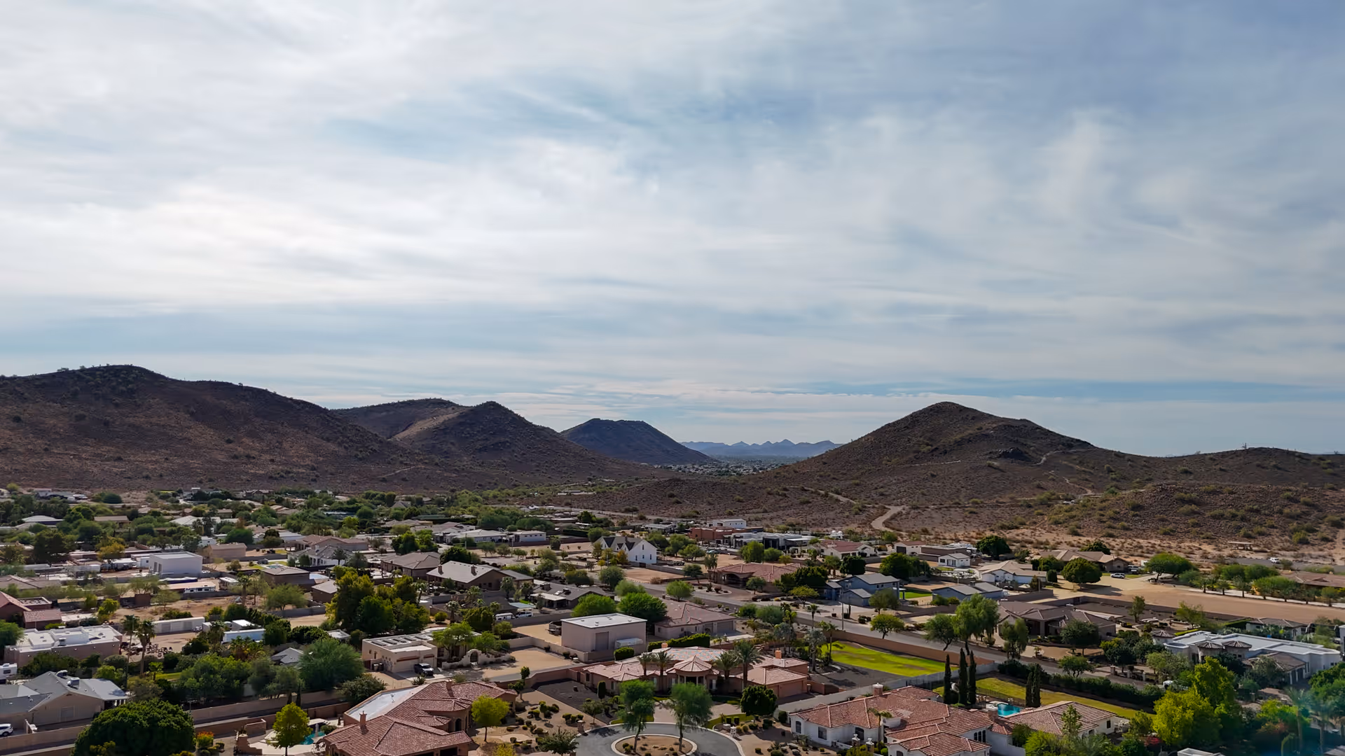 Desert suburban landscape with low houses, greenery, and distant hills under a cloudy sky.