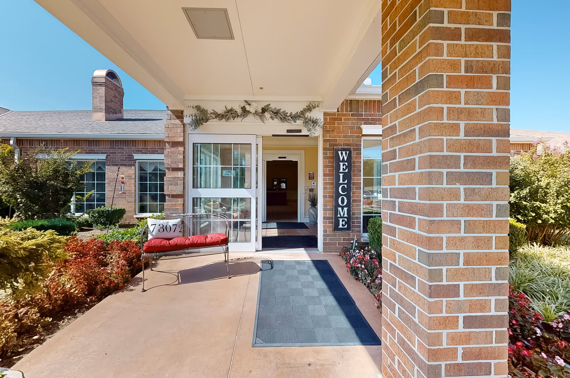 Entrance of Rivermont Assisted Living and Memory Care facility with a covered walkway, a bench with a red cushion, a welcome sign on a brick pillar, and well-maintained landscaping with bushes and flowers under a clear blue sky.
