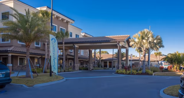 Exterior view of Inspired Living Lakewood Ranch facility showing a covered entrance with a clearance sign, palm trees, landscaped greenery, and a clear blue sky.