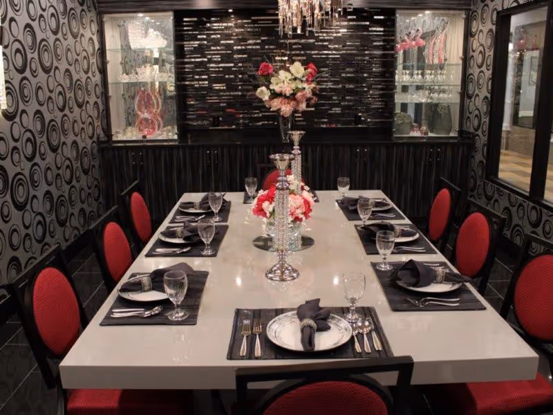 A formal dining room with a long white table set for eight people. The table is adorned with elegant silver candlesticks and floral centerpieces. Each place setting includes a plate, black napkin, silverware, and a water glass. The chairs have red upholstery and black frames. The walls have a modern black and gray circular pattern, and there are glass display cabinets with decorative items on either side of a black textured wall at the far end.